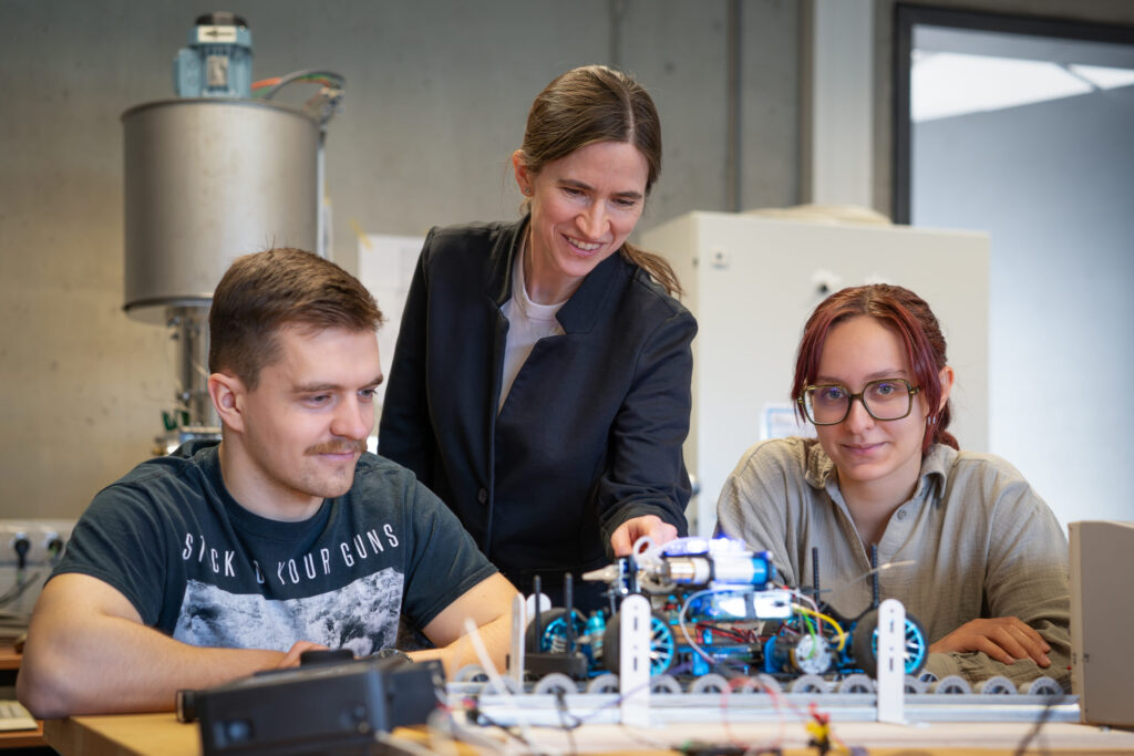 Three people work together on a robotics project in a lab. One person stands and points while the other two sit and look at the robot. They appear focused and engaged in a collaborative STEM environment. at Hochschule Coburg
