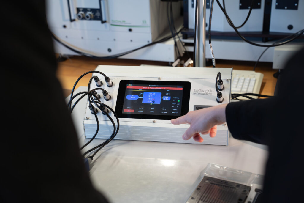 A person points at a touchscreen control panel with multiple cables connected, possibly in a laboratory or technical setting, with another person observing. at Hochschule Coburg