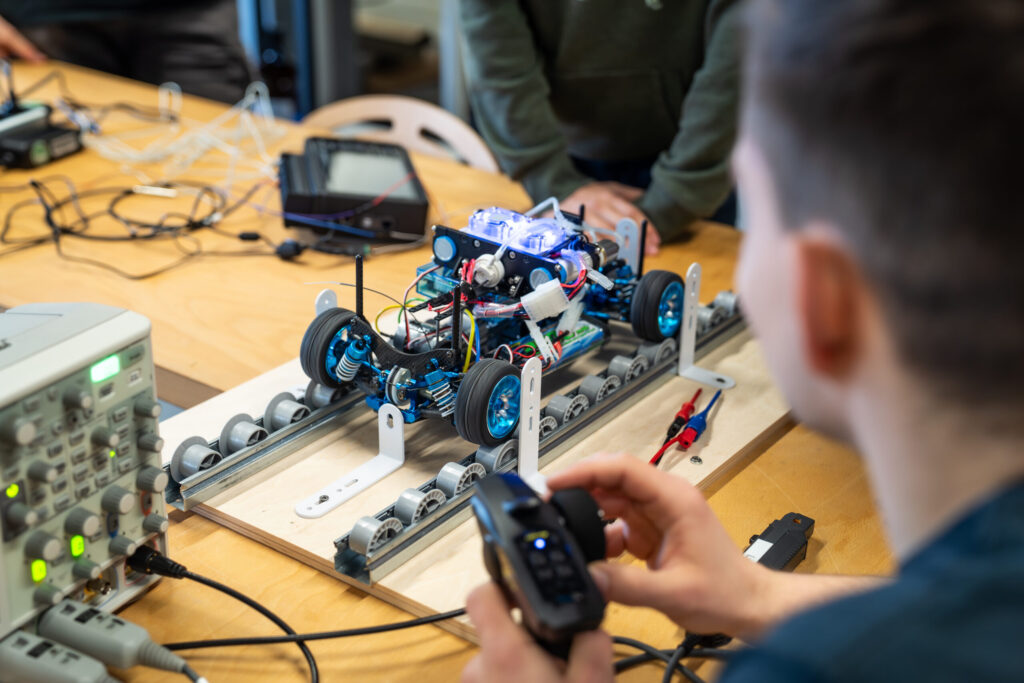 A person operates a remote-controlled car on a test track, surrounded by electronics and wires, while others observe and work in the background. at Hochschule Coburg
