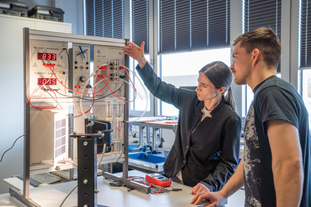 Two people stand at a lab workstation, examining and discussing a hydrogen fuel cell experiment setup with electronic displays and wires in a bright, modern classroom. at Hochschule Coburg