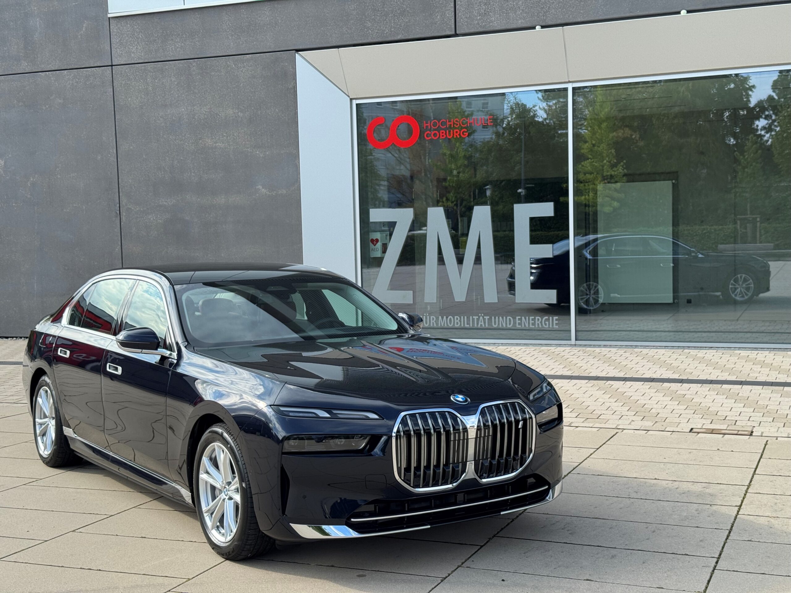 Eine schwarze BMW-Limousine steht auf einer gepflasterten Fläche vor einem modernen Gebäude mit großen Fenstern, auf denen die Buchstaben „ZME“ und ein rotes Logo zu sehen sind. at Hochschule Coburg
