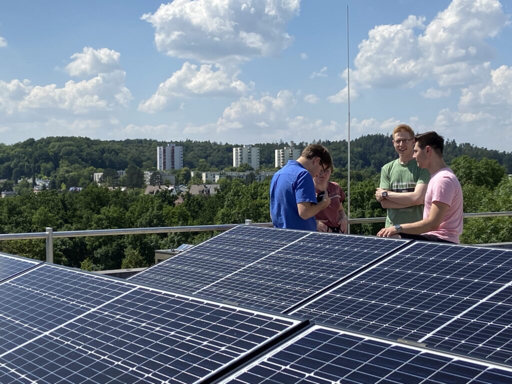 Four people stand by solar panels on a rooftop, talking and smiling, with green trees, buildings, and a partly cloudy sky in the background. at Hochschule Coburg