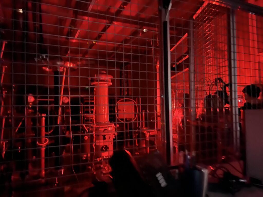 A dimly lit industrial area illuminated by red light, showing machinery behind a metal grid fence. Various pipes, valves, and equipment are visible in the enclosed space. at Hochschule Coburg