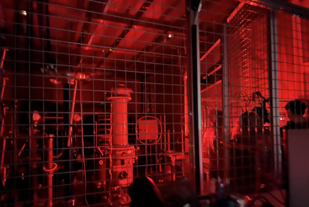 Industrieanlagen hinter einem Metallgitter werden von roten Lichtern angestrahlt, was eine dramatische und eindringliche Atmosphäre schafft. Innerhalb des umzäunten Bereichs sind Rohre, Messgeräte und Maschinen zu sehen. at Hochschule Coburg