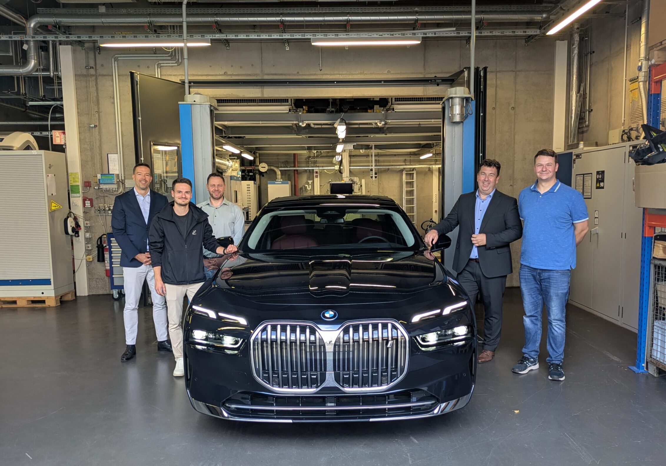 Fünf Männer stehen in einer Garage um einen schwarzen BMW herum und posieren lächelnd für das Foto. Im Hintergrund der Garage sind Regale, Werkzeuge und Industriegeräte zu sehen. at Hochschule Coburg
