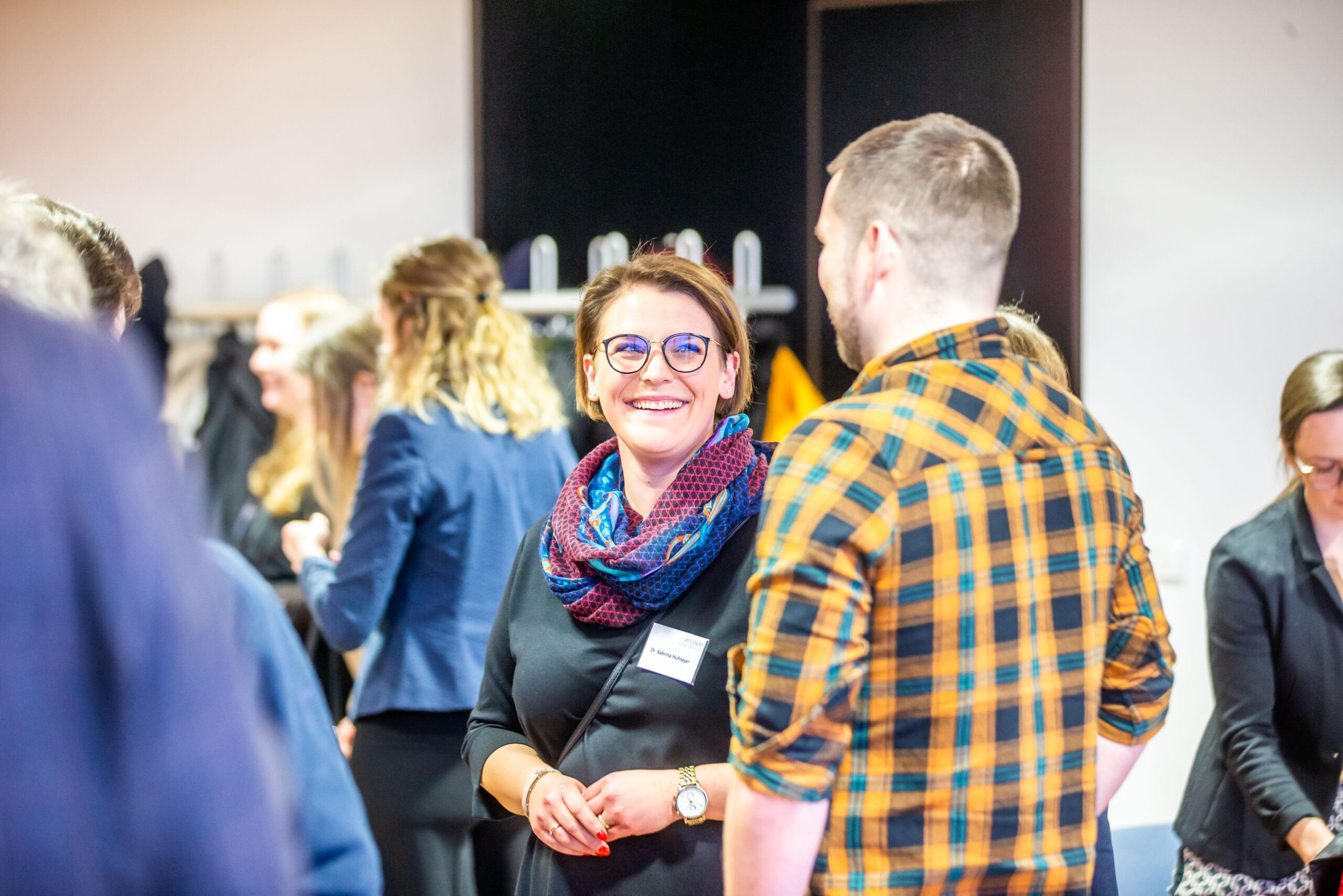 Eine Frau mit Brille und Schal lächelt, während sie sich bei einer gut besuchten Veranstaltung in einem Saal mit einem Mann in einem karierten Hemd unterhält – vielleicht sprechen sie gerade über die Möglichkeiten, die das Deutschland-Stipendium bietet. Im Hintergrund unterhalten sich weitere Personen. at Hochschule Coburg