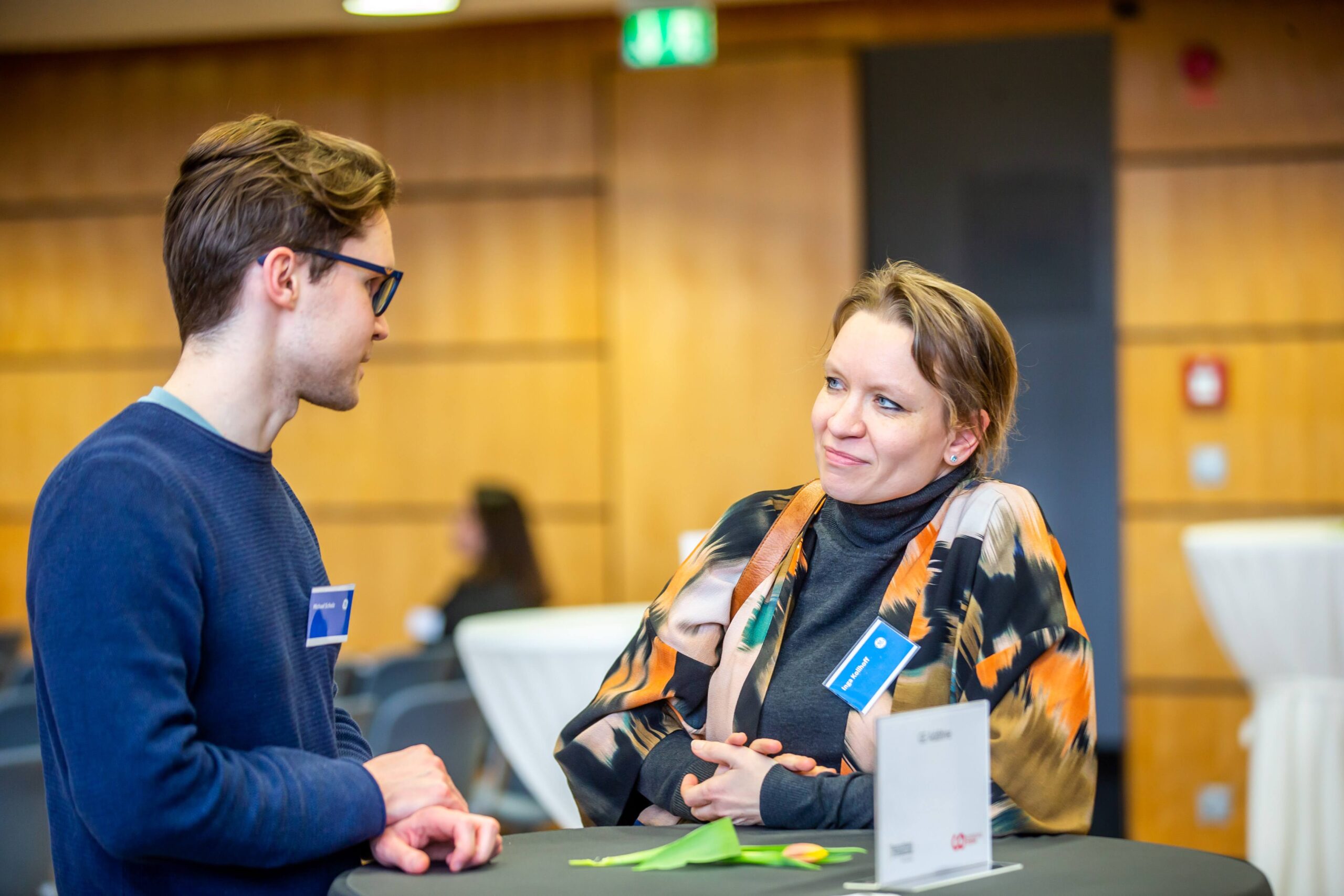 Zwei Personen stehen bei einer Veranstaltung in einem Saal an einem runden Tisch und unterhalten sich; beide tragen Namensschilder. Der Hintergrund ist unscharf, doch man erkennt Tische und andere Teilnehmer, was auf eine Networking-Veranstaltung im Rahmen eines Förderprogramms wie dem Deutschland-Stipendium hindeutet. at Hochschule Coburg