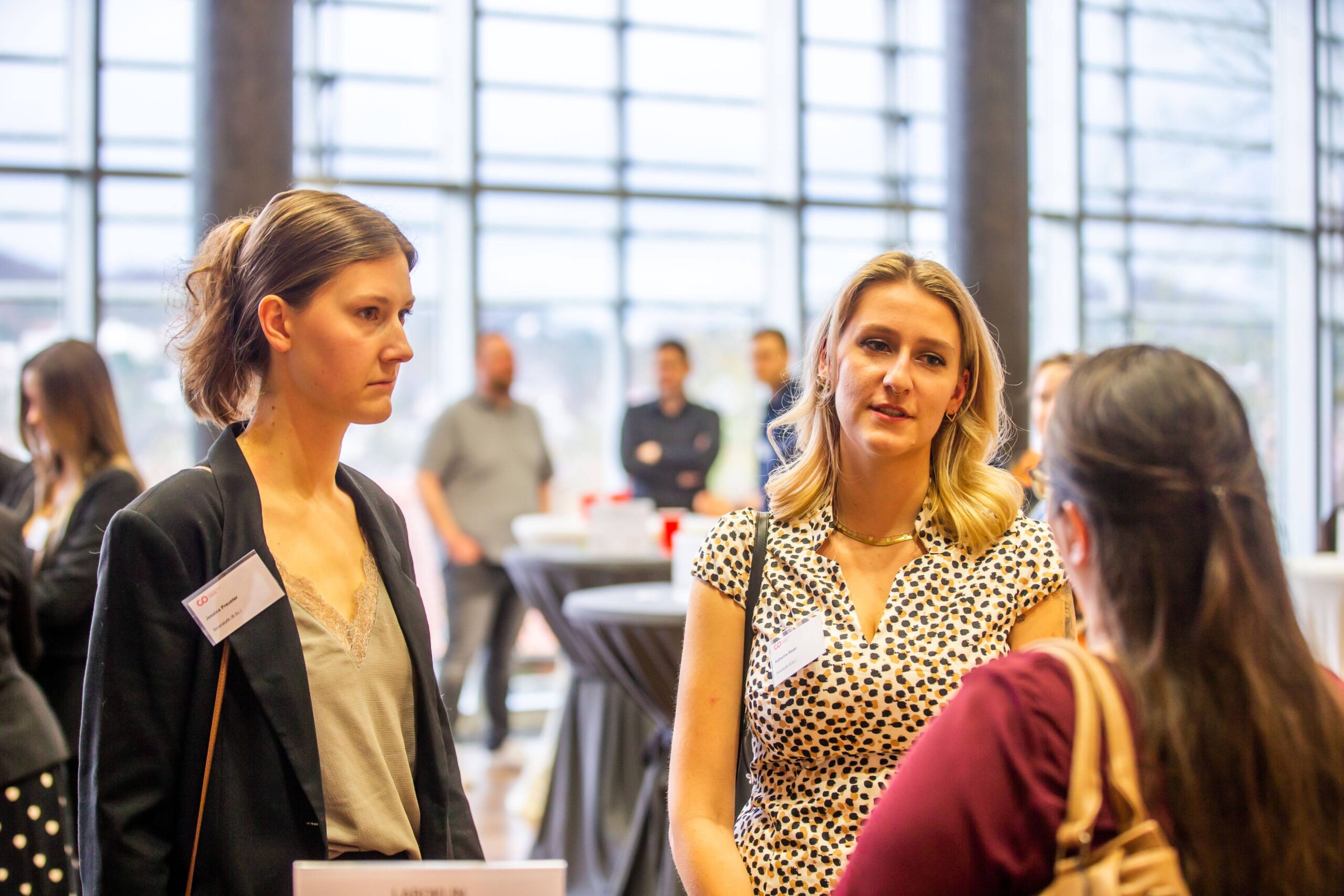 Drei Frauen stehen bei einer Networking-Veranstaltung in einem Raum und unterhalten sich, während im unscharfen Hintergrund weitere Personen und Cocktailtische zu sehen sind. Zwei Frauen blicken in die Kamera, während eine den Rücken zur Kamera gewandt hat. Sie sprechen über neue Chancen und das Potenzial, das sich durch den Erhalt eines Deutschland-Stipendiums eröffnet. at Hochschule Coburg