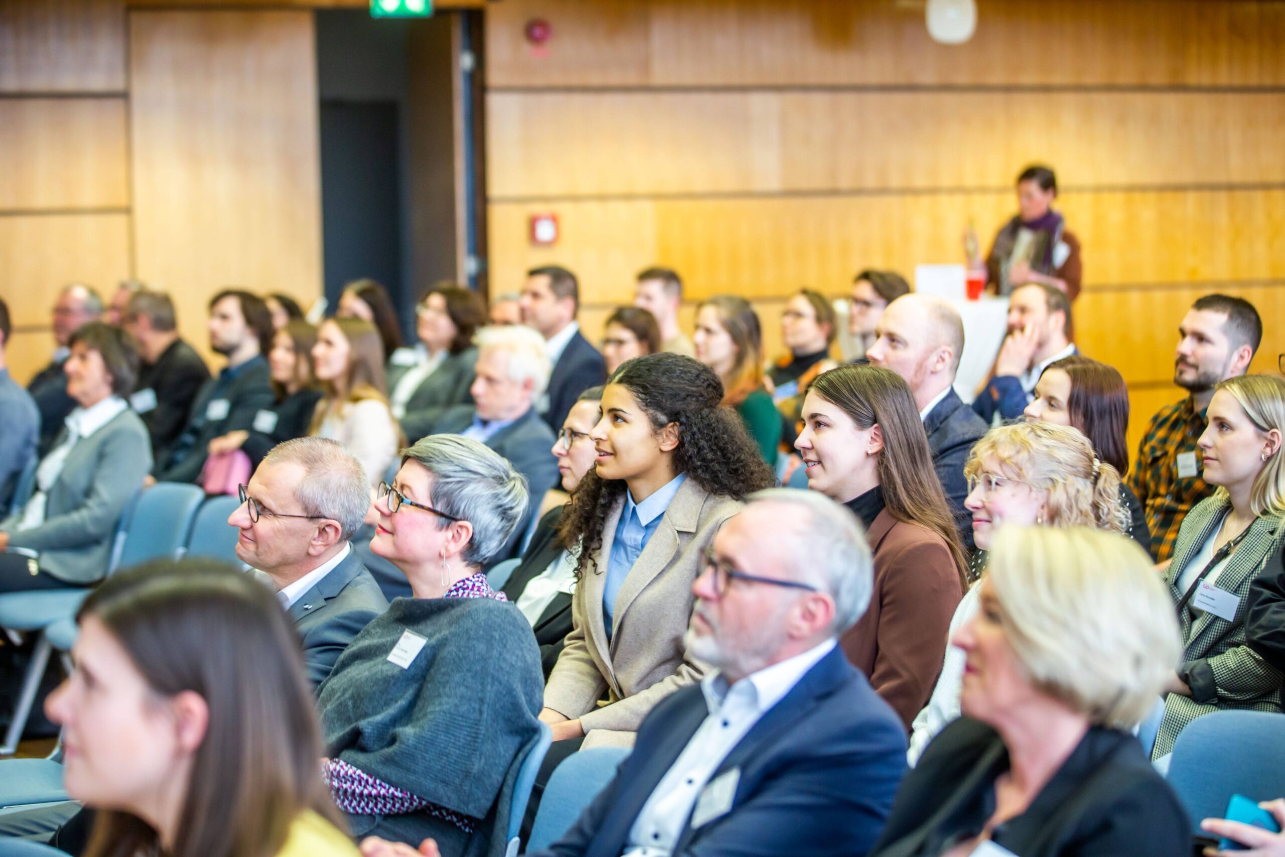 Teilnehmer mit großem Potenzial sitzen bei einer Konferenz oder einem Seminar in einem Saal aufmerksam in Reihen und blicken nach vorne. Anwesend sind sowohl Männer als auch Frauen unterschiedlichen Alters, die sich möglicherweise versammelt haben, um mehr über Fördermöglichkeiten wie das Deutschland-Stipendium zu erfahren. Im Hintergrund sind helle Holzvertäfelungen zu sehen sowie eine Person, die weiter hinten steht. at Hochschule Coburg