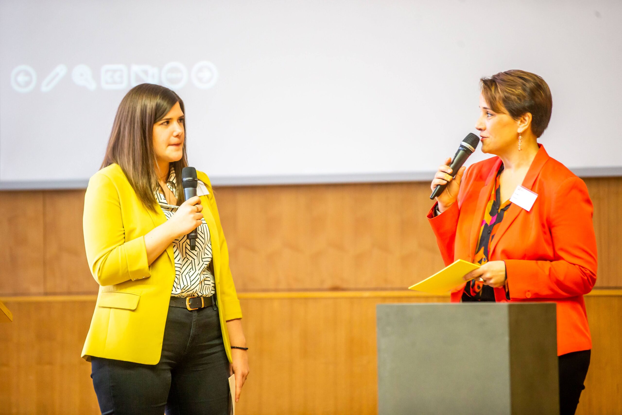Zwei Frauen stehen sich gegenüber, jede hält ein Mikrofon in der Hand. Die eine trägt einen gelben Blazer, die andere einen roten, und sie scheinen bei einer Veranstaltung oder Präsentation in einem Saal zu sprechen, vielleicht um über Fördermöglichkeiten wie das Deutschland-Stipendium zu informieren und andere zu ermutigen, ihr Potenzial durch dieses renommierte Stipendium zu entfalten. at Hochschule Coburg
