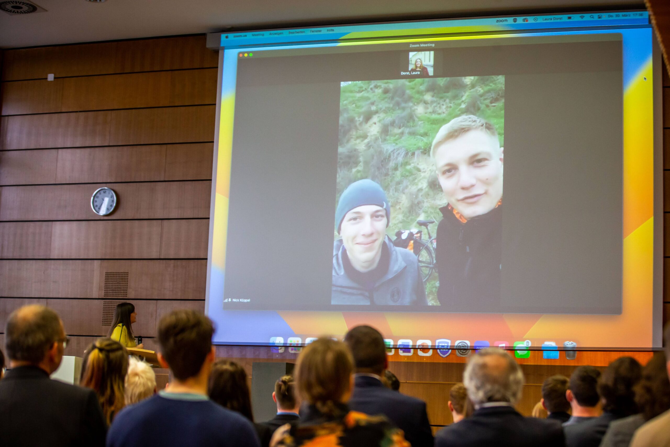 Eine Gruppe von Menschen in einem Auditorium schaut auf eine große Leinwand, auf der ein Videoanruf mit zwei lächelnden Personen im Freien zu sehen ist, die vielleicht über ihre Erfahrungen als Empfänger des Deutschland-Stipendiums berichten. Eine Frau steht am Rednerpult vor dem Publikum und spricht über Potenzial und Chancen. at Hochschule Coburg
