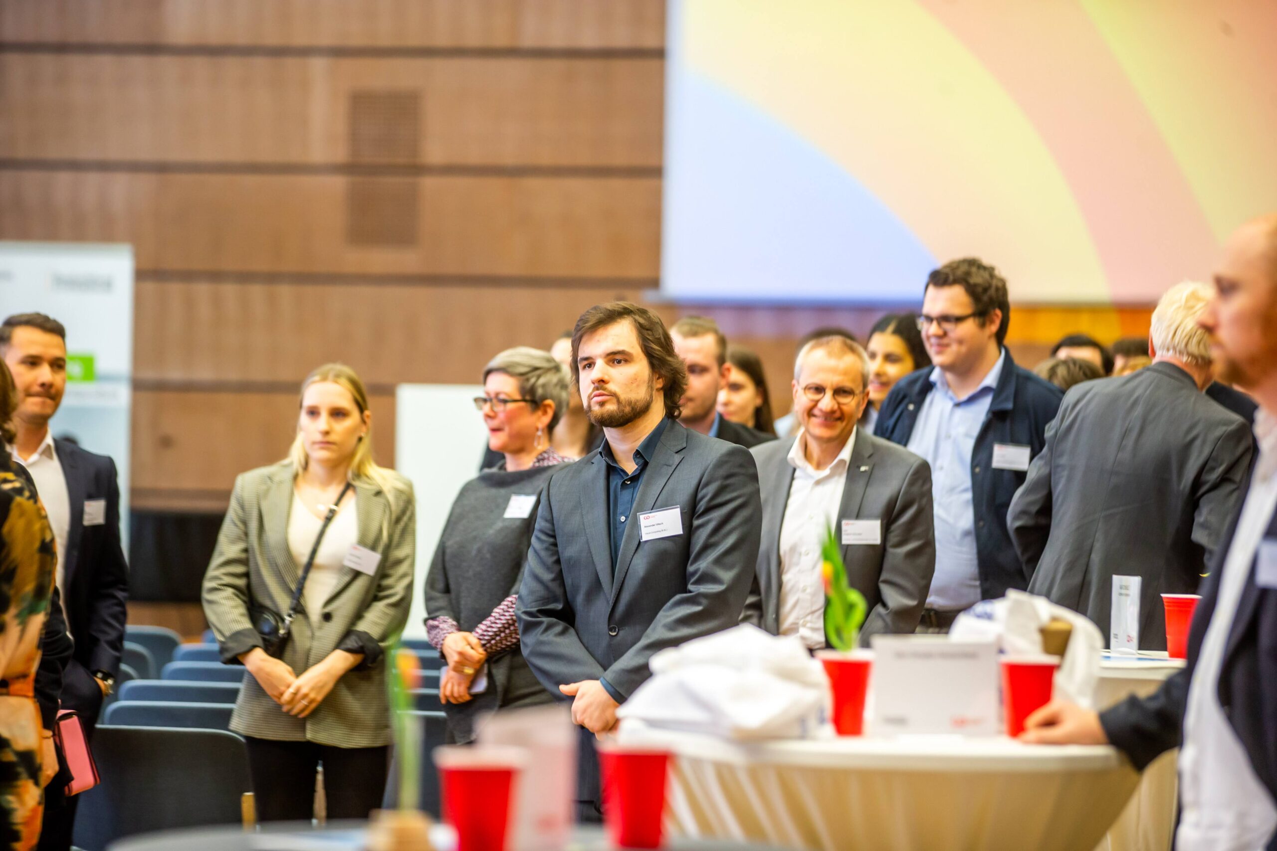 Eine Gruppe professionell gekleideter Menschen steht und unterhält sich bei einer Veranstaltung des Deutschland-Stipendiums in einem Saal, in dem runde Tische, rote Becher und Namensschilder zu sehen sind. Im Hintergrund sind eine Projektionsfläche und unscharfer Text zu erkennen, die die Würdigung akademischen Potenzials und den Wert eines Stipendiums hervorheben. at Hochschule Coburg