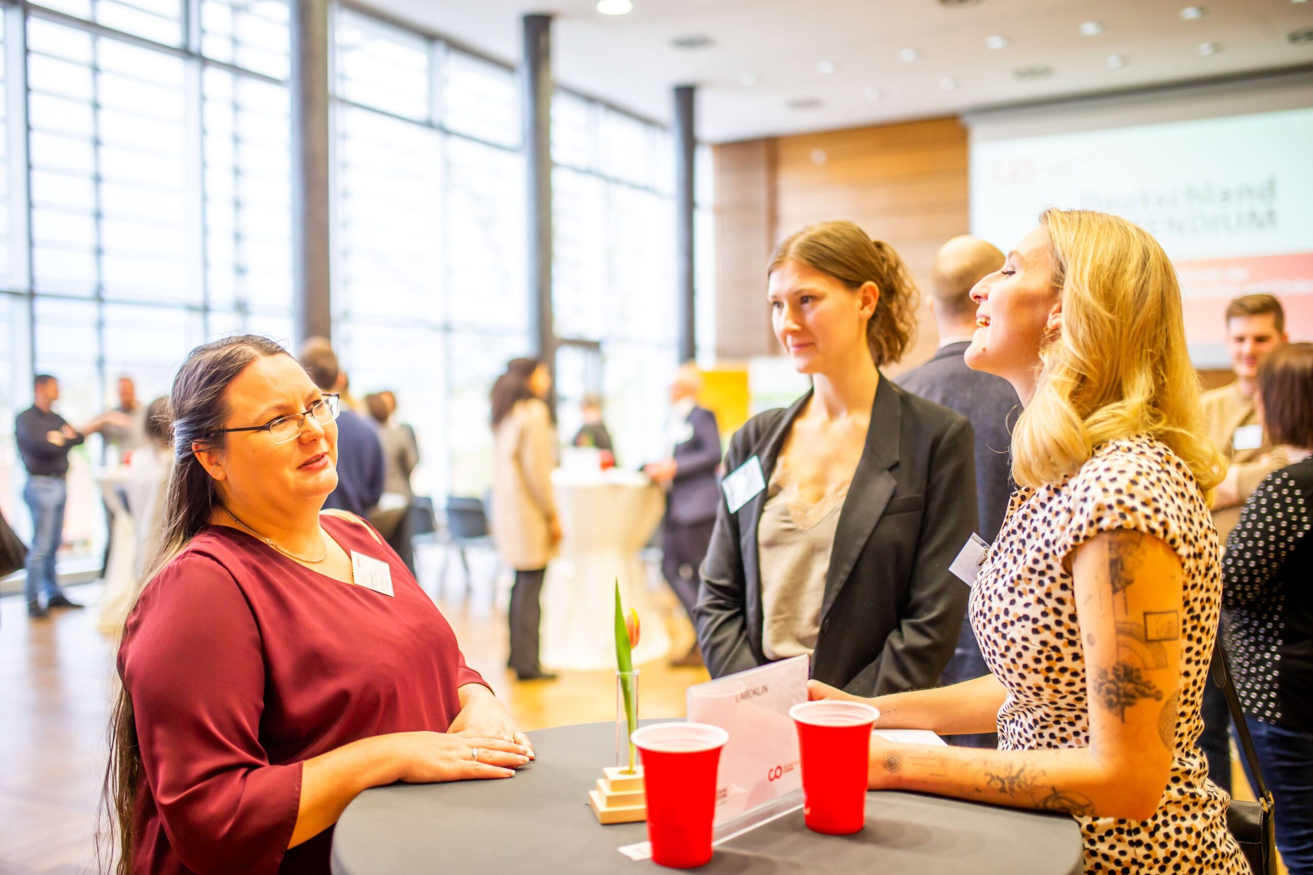 Drei Frauen stehen mit roten Bechern um einen hohen Tisch herum und unterhalten sich bei einer hell beleuchteten, modernen Networking-Veranstaltung über ihr Potenzial. Im Hintergrund unterhalten sich andere Gruppen, vielleicht tauschen sie Erfahrungen zum Deutschland-Stipendium aus. An der Wand ist eine unscharfe Präsentationsfläche zu sehen. at Hochschule Coburg