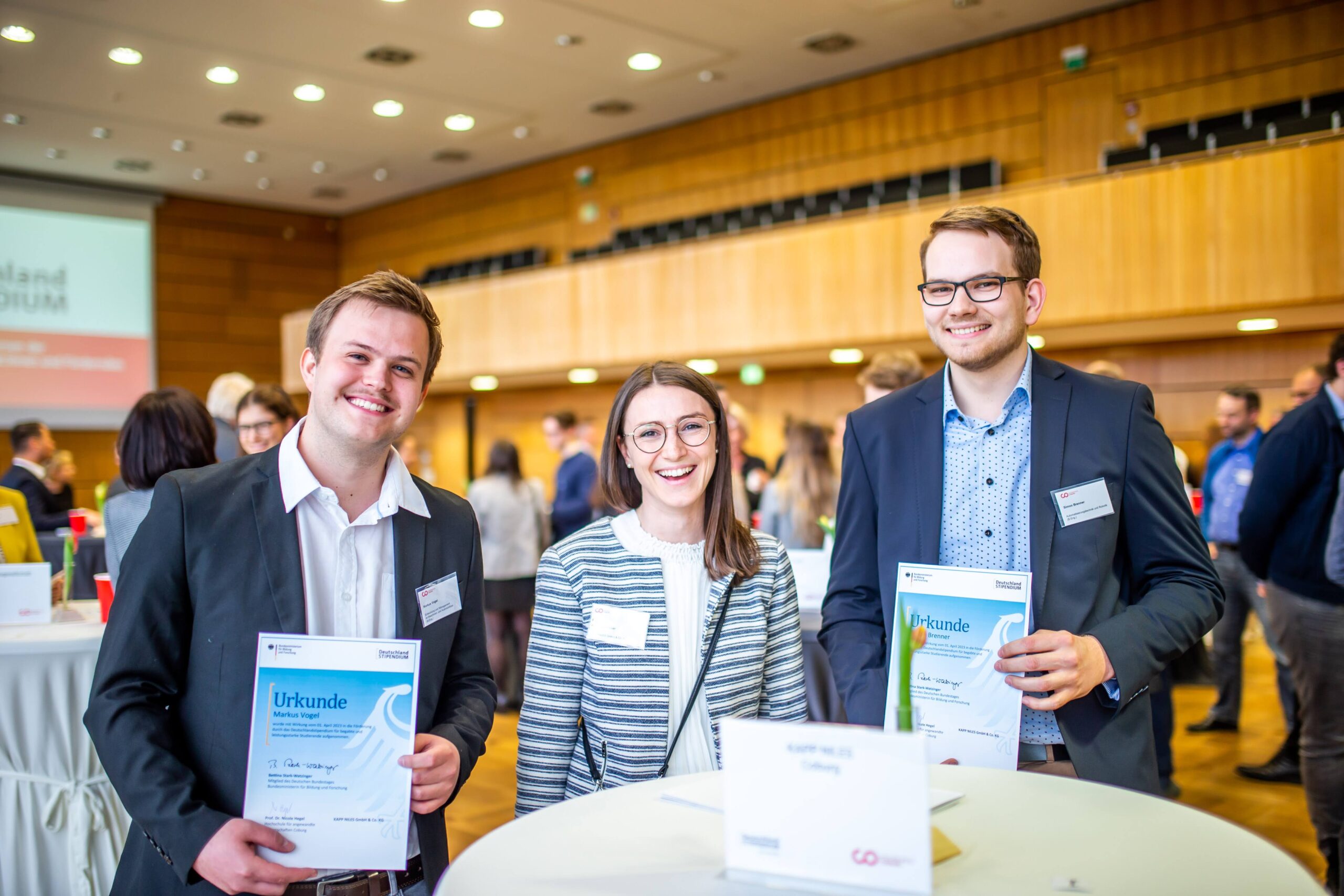 Drei lächelnde junge Erwachsene in Business-Kleidung stehen an einem runden Tisch und halten jeweils stolz eine Urkunde des Deutschland-Stipendiums in den Händen. Im Hintergrund füllen weitere Menschen und Konferenztische den hellen, modernen Veranstaltungssaal und unterstreichen die feierliche Atmosphäre dieses renommierten Förderprogramms. at Hochschule Coburg
