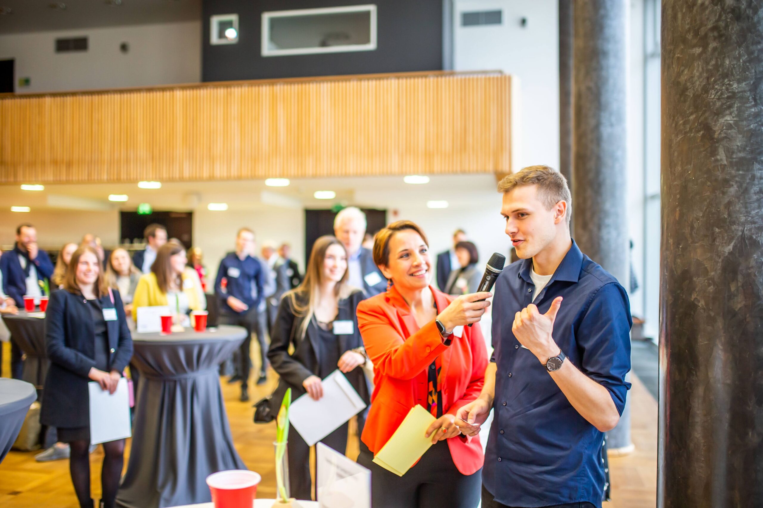 Eine Frau in einem roten Blazer interviewt bei einer Networking-Veranstaltung einen Mann mit einem Mikrofon, während sich im Hintergrund Menschen um hohe Tische versammeln, sich unterhalten und über „Potenzial“ sowie Möglichkeiten wie das Deutschland-Stipendium sprechen. at Hochschule Coburg