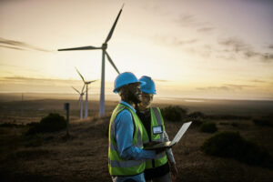 Two workers wearing safety vests and helmets look at a laptop at sunset in a wind farm; wind turbines are visible in the background.