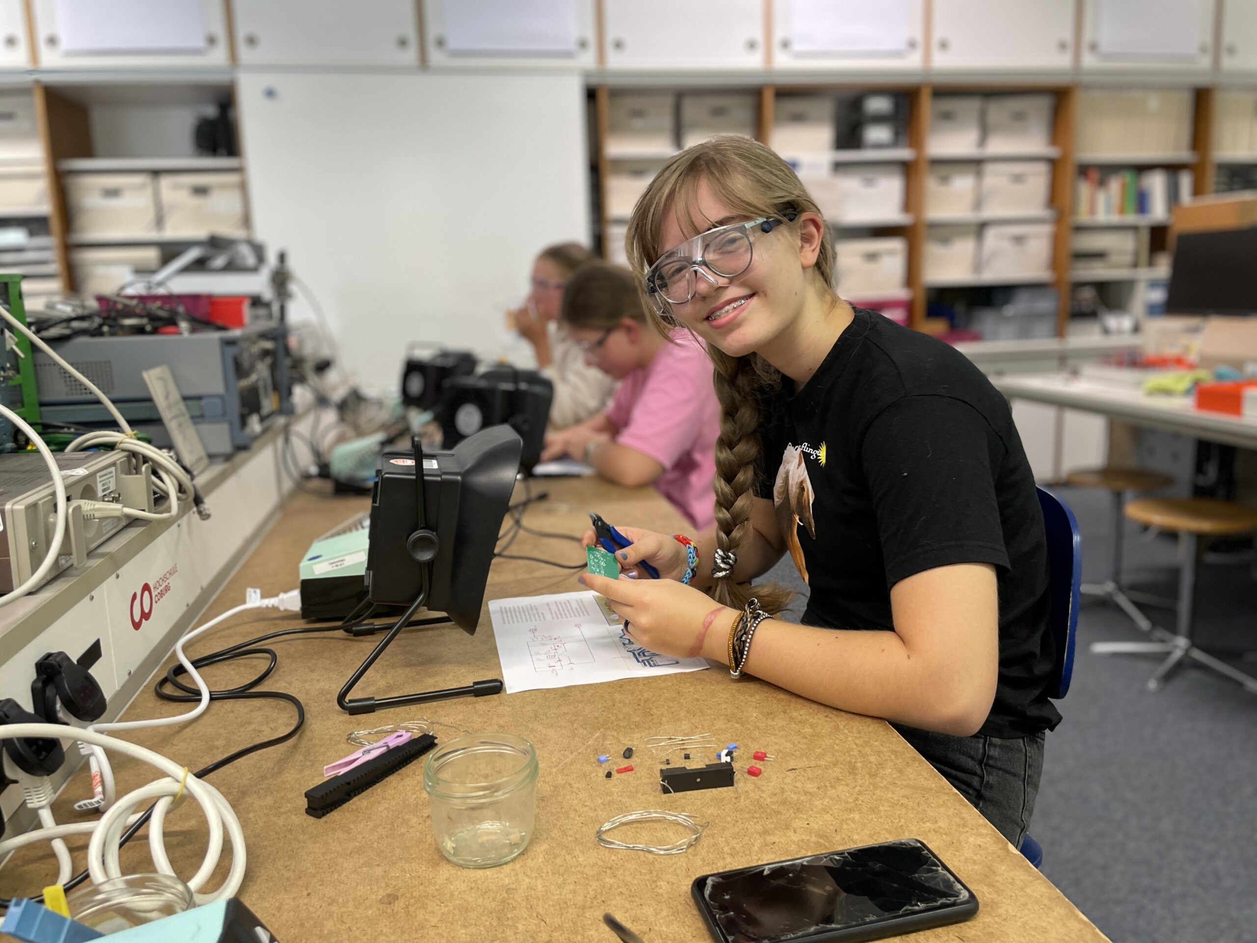 Ein Mädchen mit Schutzbrille lächelt, während es im Rahmen der MUT-Ferienaktion an einem Labortisch mit Elektronik arbeitet und dabei neue Möglichkeiten in der Technik entdeckt. Im Hintergrund sind zwei weitere Personen bei der Arbeit zu sehen. Auf dem Tisch liegen verschiedene elektronische Bauteile und Geräte. at Hochschule Coburg