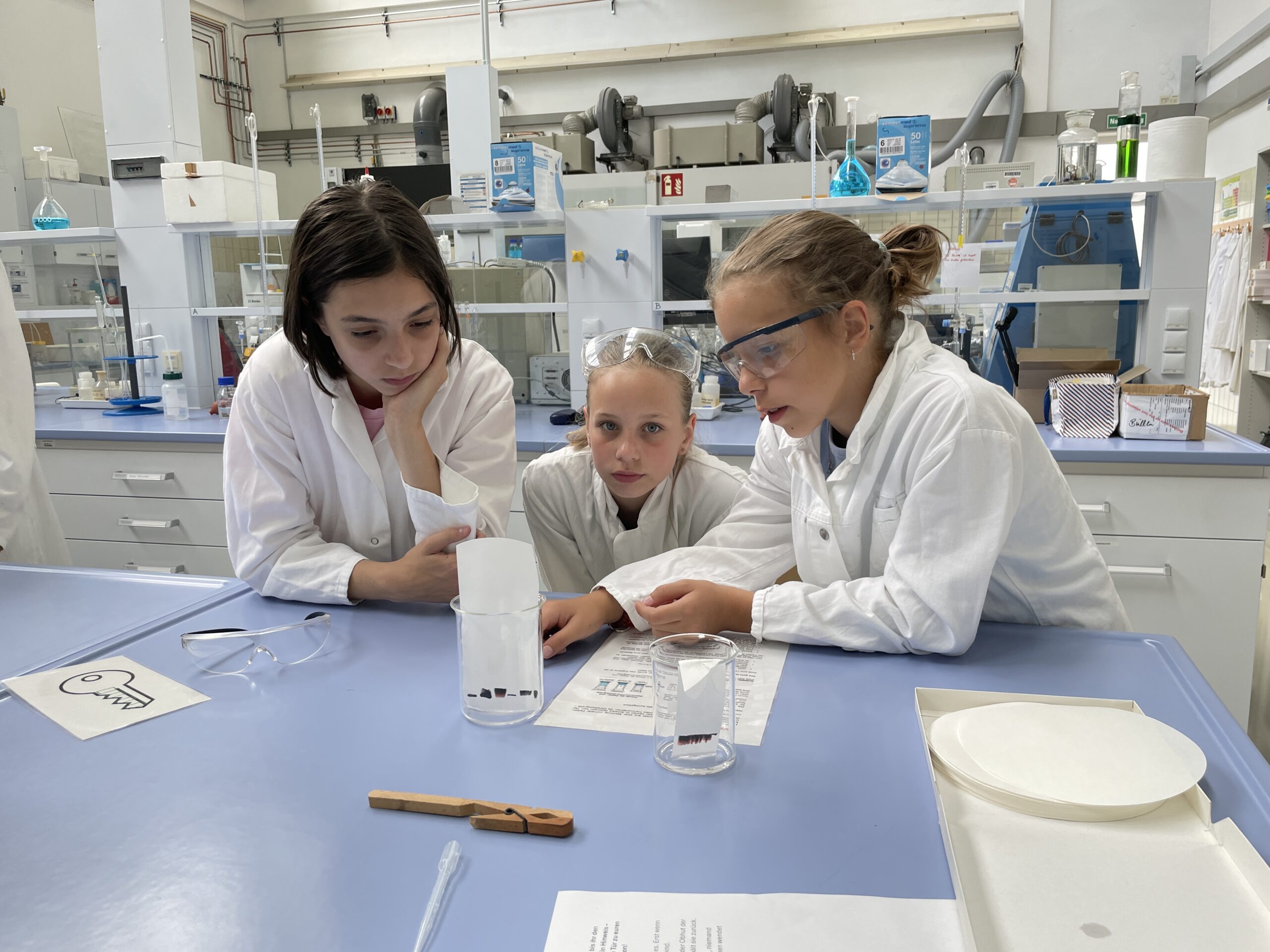 Drei Mädchen in Laborkitteln und Schutzbrillen untersuchen ein Becherglas und ein Blatt Papier auf einem blauen Labortisch; sie wirken konzentriert und sind ganz in ein wissenschaftliches Experiment im Rahmen der MUT-Ferienaktion vertieft. Im Hintergrund ist Laborausrüstung zu sehen, was verdeutlicht, wie die Teilnehmerinnen durch praktisches Ausprobieren neue Möglichkeiten entdecken. at Hochschule Coburg