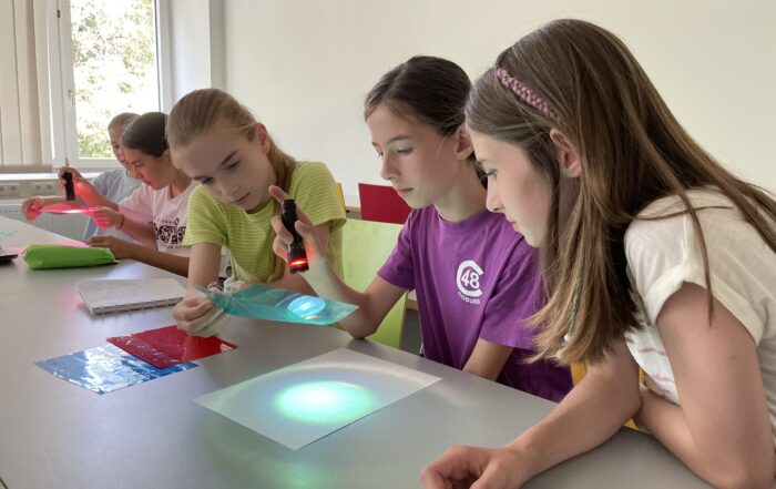 Vier Mädchen in einem Klassenzimmer erkunden mit Taschenlampen und farbigen Transparentfolien Licht und Farbe auf einer weißen Fläche und entdecken im Rahmen der MUT-Ferienaktion durch ihr praktisches Experiment neue Möglichkeiten. at Hochschule Coburg