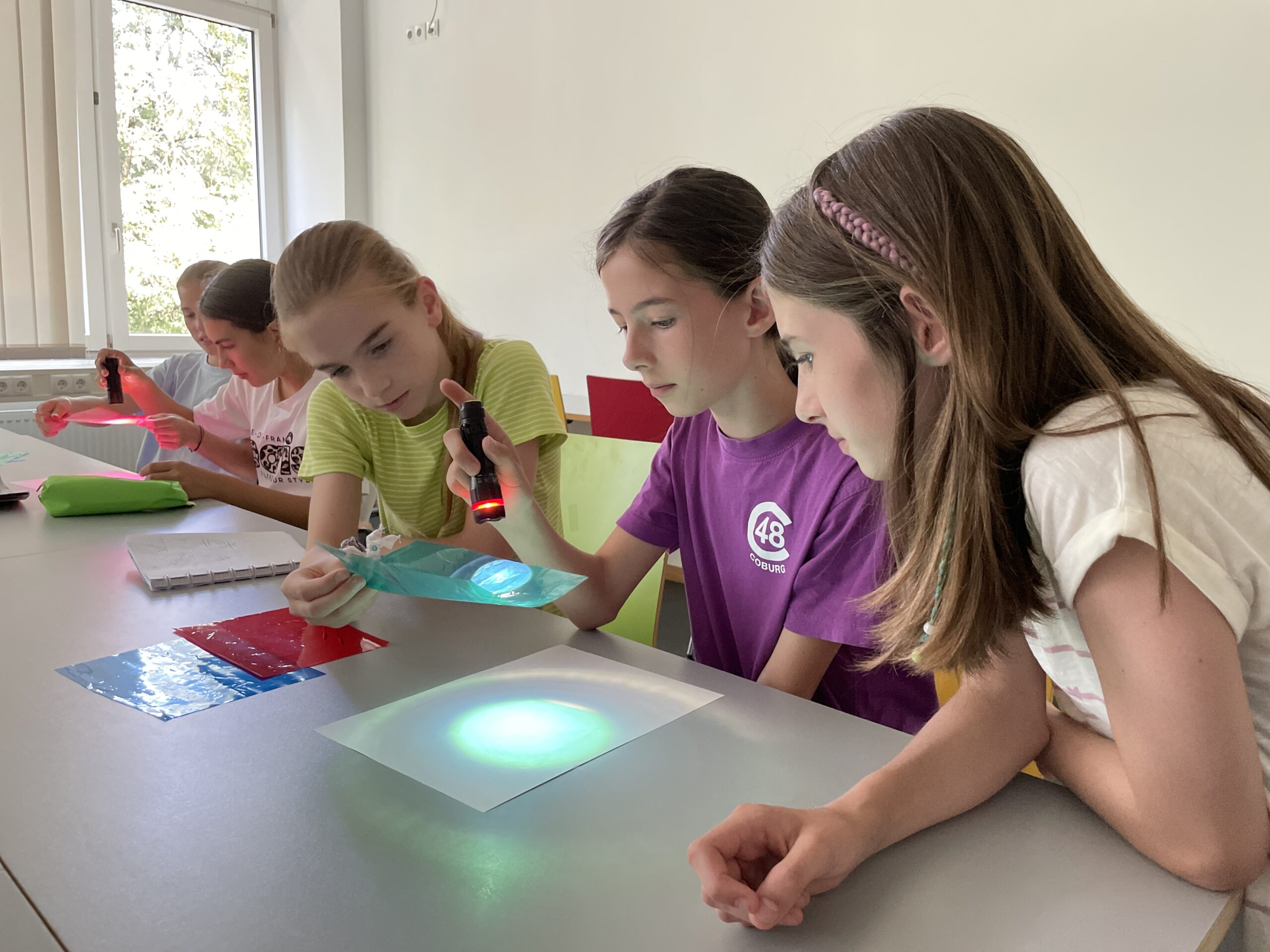 Vier Mädchen in einem Klassenzimmer erkunden mit Taschenlampen und farbigen Transparentfolien Licht und Farbe auf einer weißen Fläche und entdecken im Rahmen der MUT-Ferienaktion durch ihr praktisches Experiment neue Möglichkeiten. at Hochschule Coburg