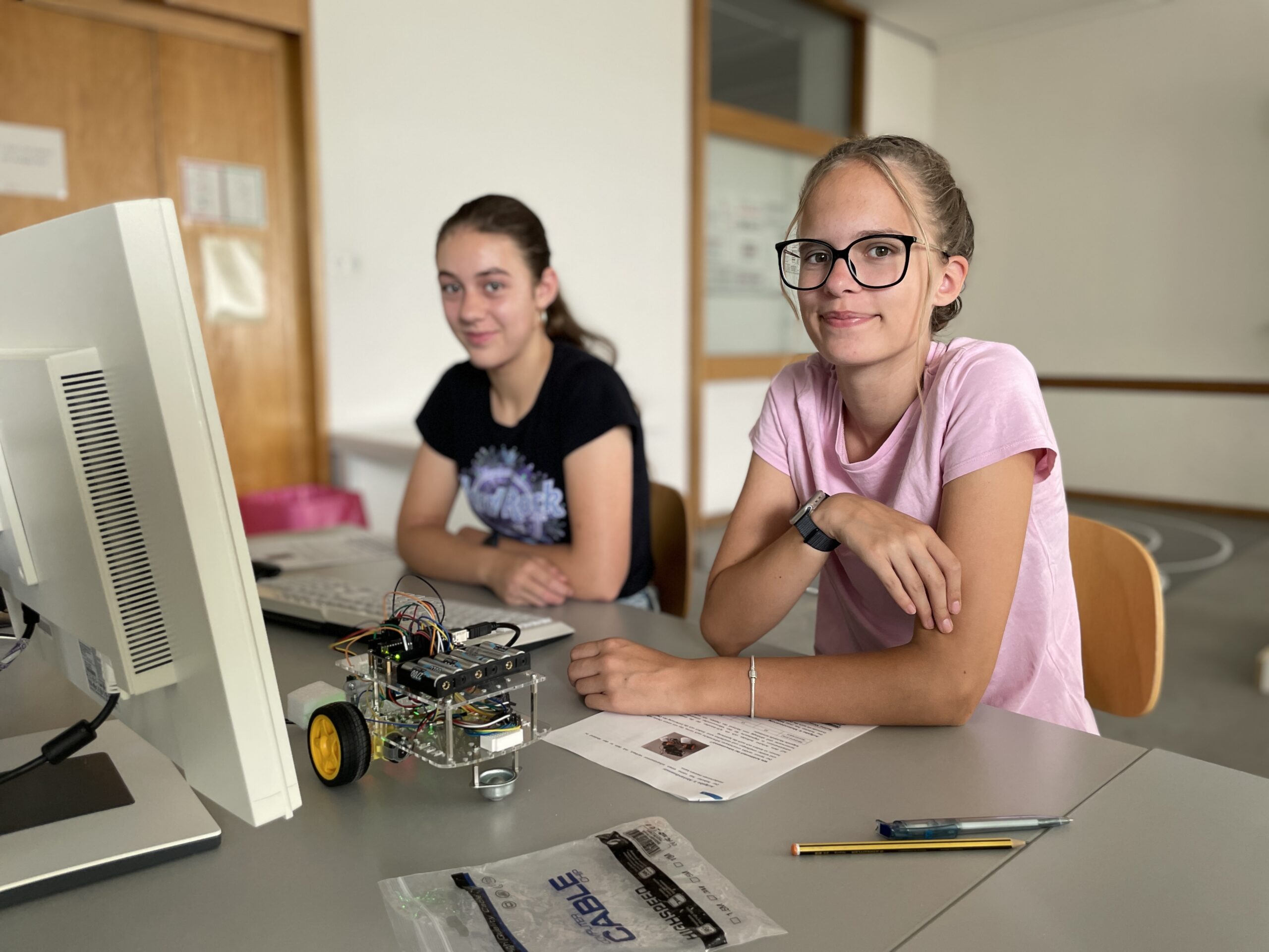 Zwei Mädchen sitzen an einem Schreibtisch mit einem Computer und einem kleinen Roboter auf Rädern und nehmen an der MUT-Ferienaktion teil. Die eine trägt eine Brille und ein rosa Shirt, die andere ein schwarzes Shirt. Umgeben von Papieren und Stiften in einem hellen Klassenzimmer erkunden sie gemeinsam neue Möglichkeiten in den Bereichen Technik und Robotik. at Hochschule Coburg