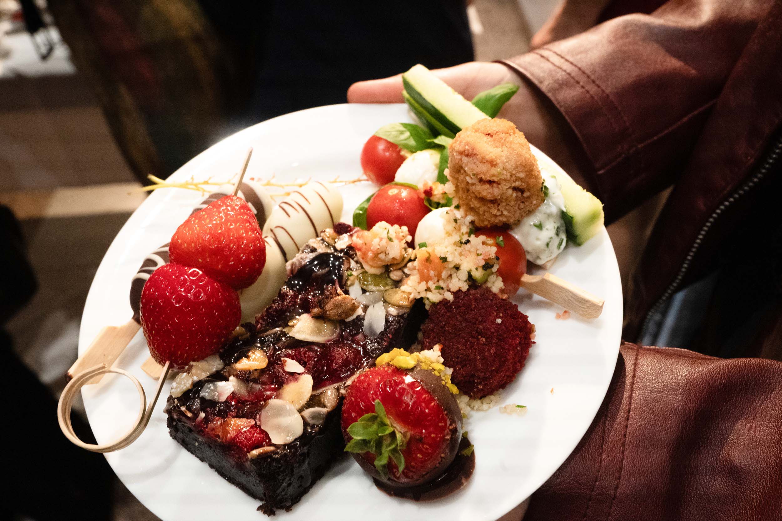 Eine Hand hält einen weißen Teller mit verschiedenen Häppchen und Desserts, darunter Erdbeeren, in Schokolade getauchte Leckereien, einen Brownie mit Mandeln, Tomaten und einen Falafel-Spieß – perfekt, um den Abschied von Absolventinnen und Absolventen zu feiern. at Hochschule Coburg