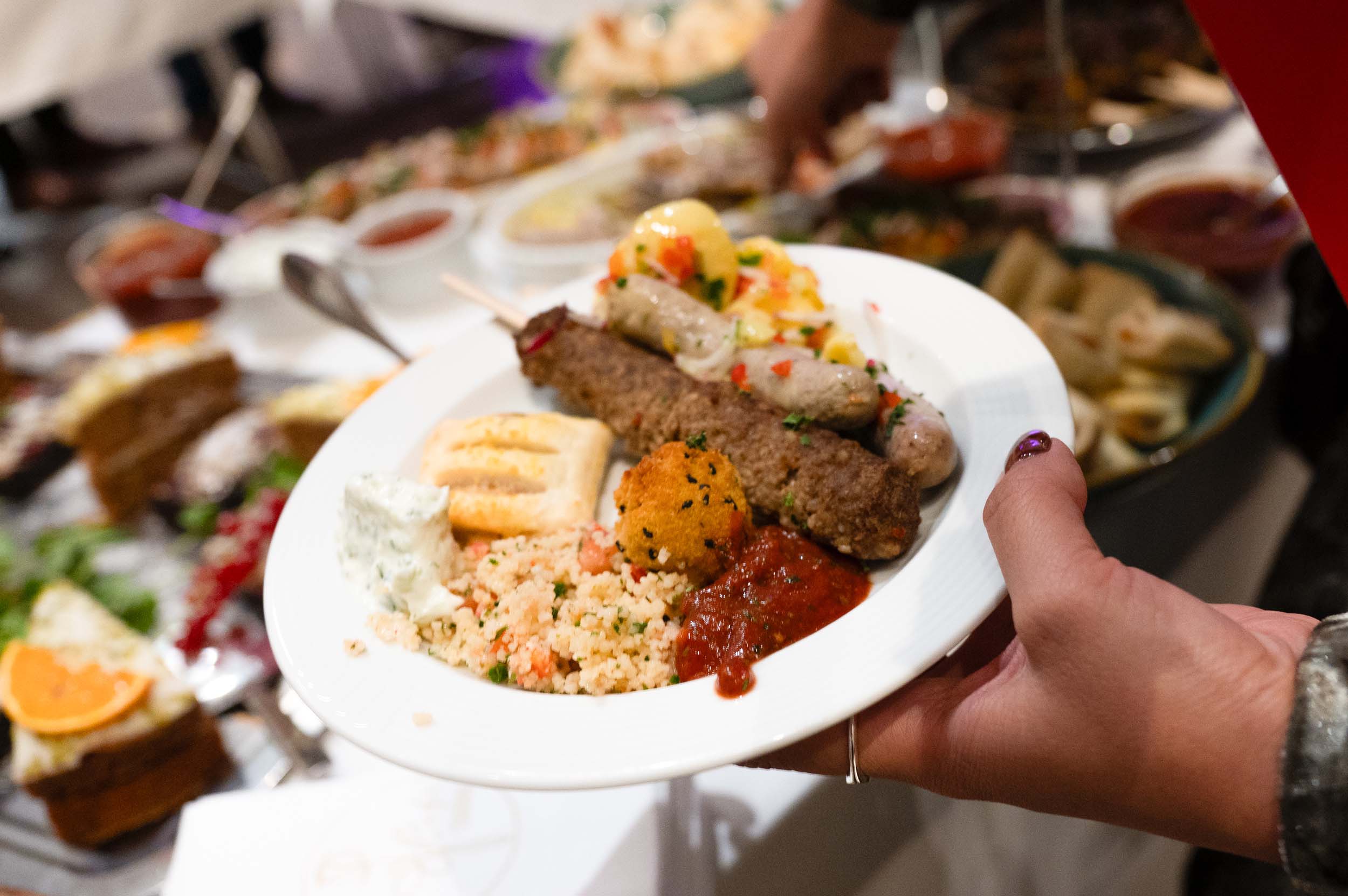 Eine Hand hält einen Teller mit Kebab, Brot, Taboulé, roter Soße, einer Falafel und Tzatziki – eine verlockende Auswahl, die sich perfekt für einen Abschied oder eine Feier zu Ehren von Absolventen und Absolventinnen eignet. Im Hintergrund sind weitere verschiedene Gerichte nur schemenhaft zu erkennen, was auf ein festliches Buffet hindeutet. at Hochschule Coburg