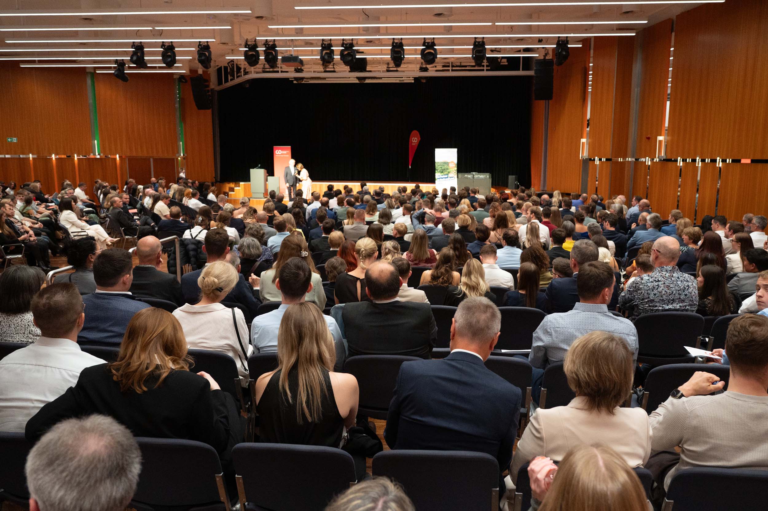 Ein großes Publikum, darunter Absolventen und Absolventinnen, sitzt in Reihen vor einer Bühne, auf der ein Redner in einem geräumigen, modernen Hörsaal mit holzvertäfelten Wänden und Deckenleuchten eine Rede hält – ein unvergesslicher Abschied. at Hochschule Coburg