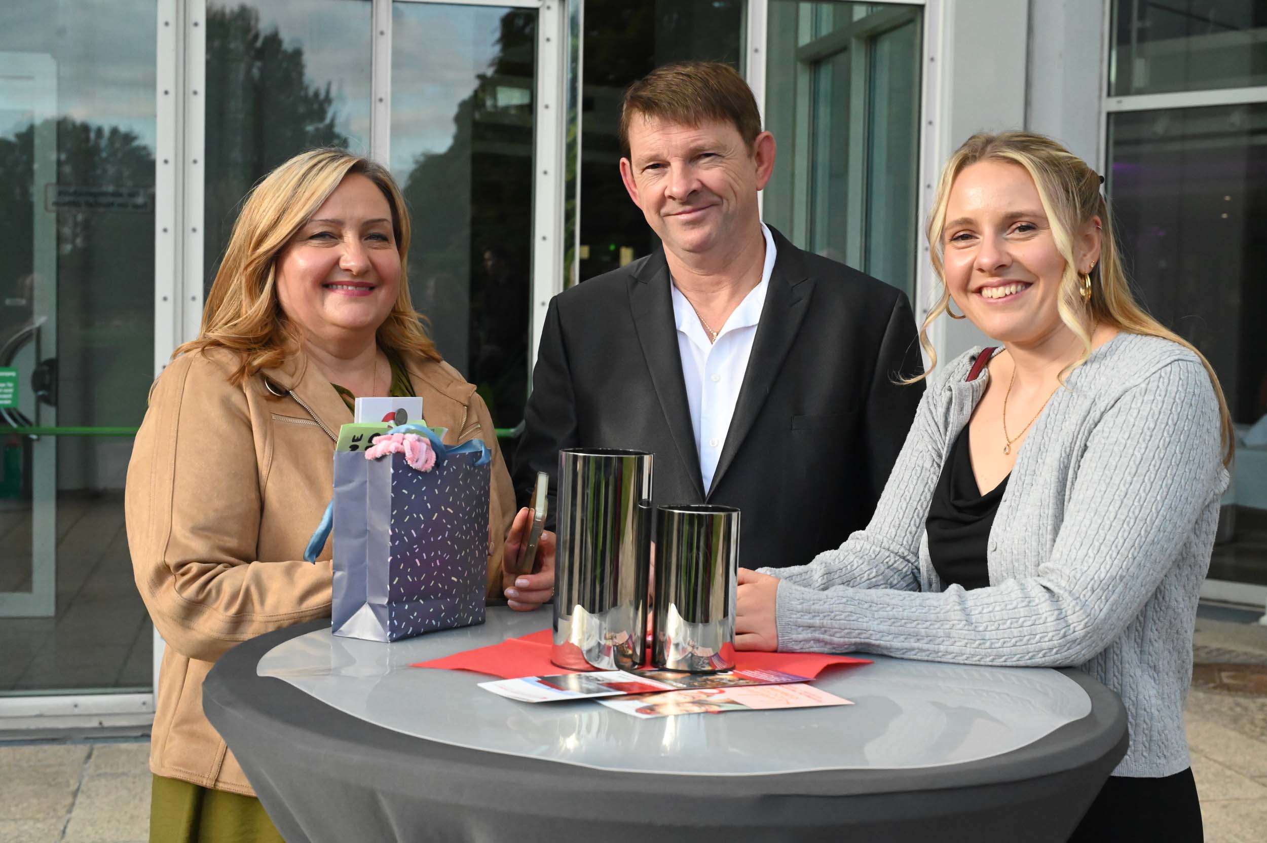 Drei Personen stehen im Freien um einen runden Tisch herum und lächeln in die Kamera – vielleicht feiern sie einen Abschied. Auf dem Tisch stehen zwei hohe Silberbecher, eine gemusterte Geschenktüte sowie einige Karten oder Broschüren. Im Hintergrund sind Glastüren und Bäume zu sehen. at Hochschule Coburg