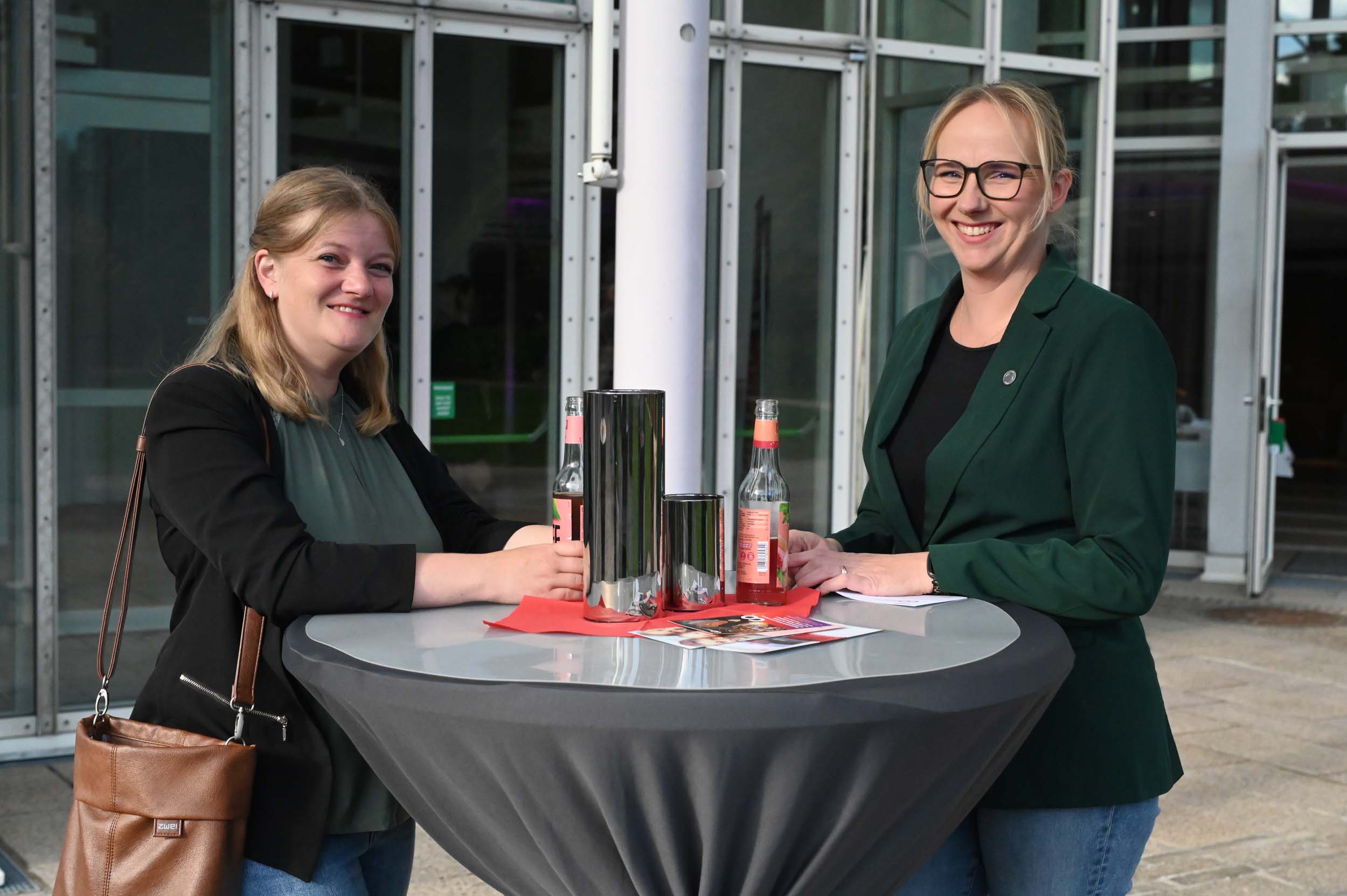 Zwei Frauen stehen an einem runden Tisch mit Getränken und Papieren und lächeln in die Kamera. Beide tragen dunkle Blazer und eine Brille. Sie befinden sich draußen in der Nähe von Glastüren und feiern vielleicht ihren Abschied als Absolventinnen. at Hochschule Coburg