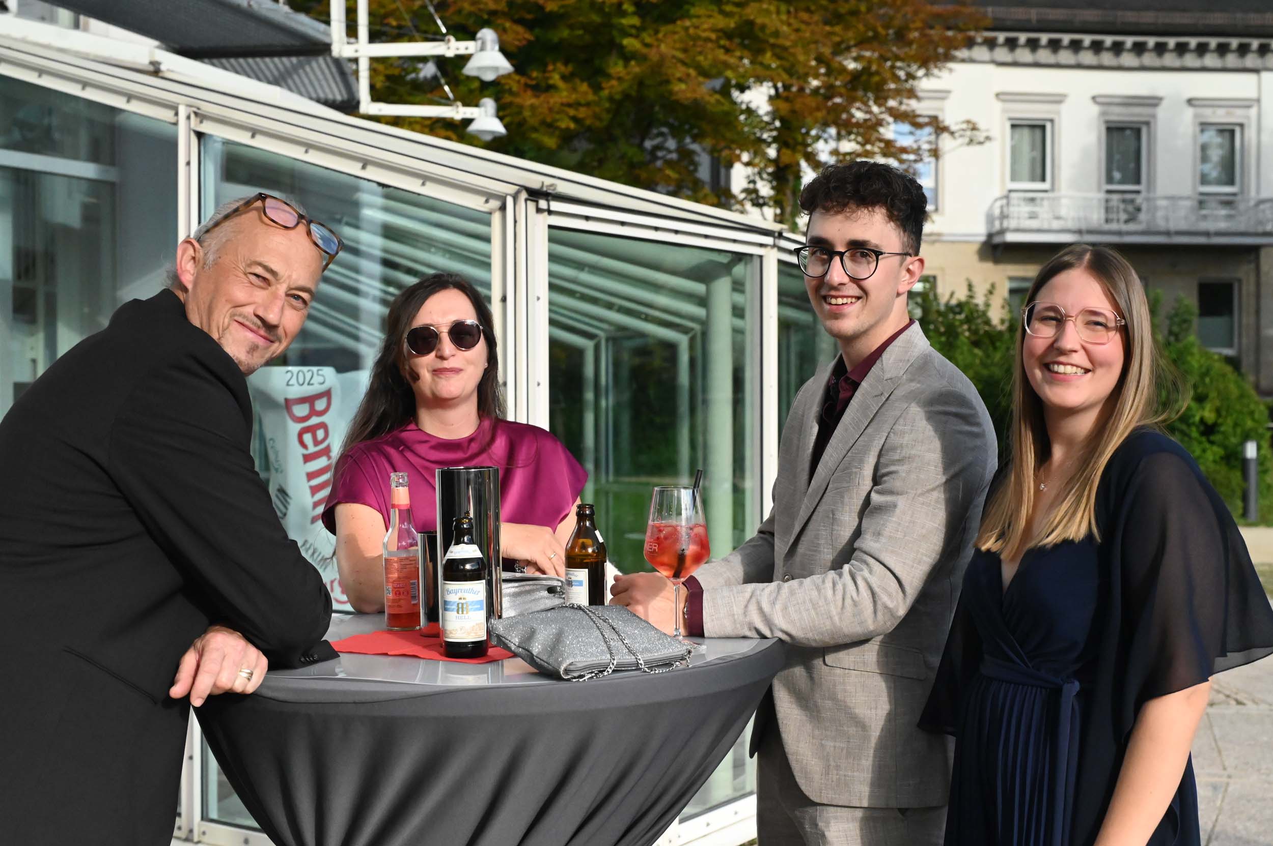 Vier festlich gekleidete Absolventinnen und Absolventen stehen im Freien um einen Cocktailtisch herum und lächeln in die Kamera. Auf dem Tisch stehen Flaschen, Gläser und eine kleine Handtasche, im Hintergrund sind Gebäude und Bäume zu sehen – ein fröhlicher Abschiedsmoment. at Hochschule Coburg