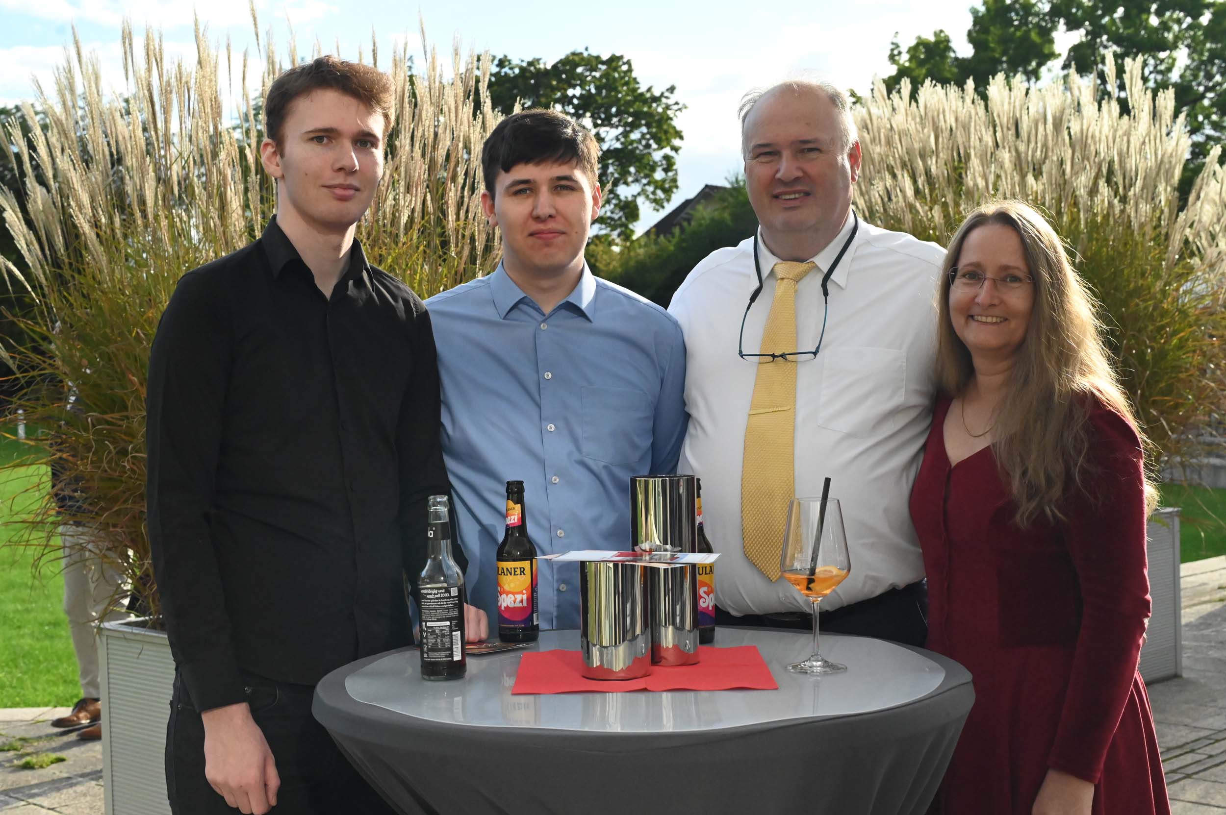 Vier Personen stehen draußen an einem runden Tisch mit Getränken und Flaschen und lächeln in die Kamera, während sie ihren Abschied feiern. Im Hintergrund sind an diesem sonnigen Tag hohes Gras und ein grüner Rasen zu sehen – ein freudiger Moment für diese Absolventen und Absolventinnen. at Hochschule Coburg
