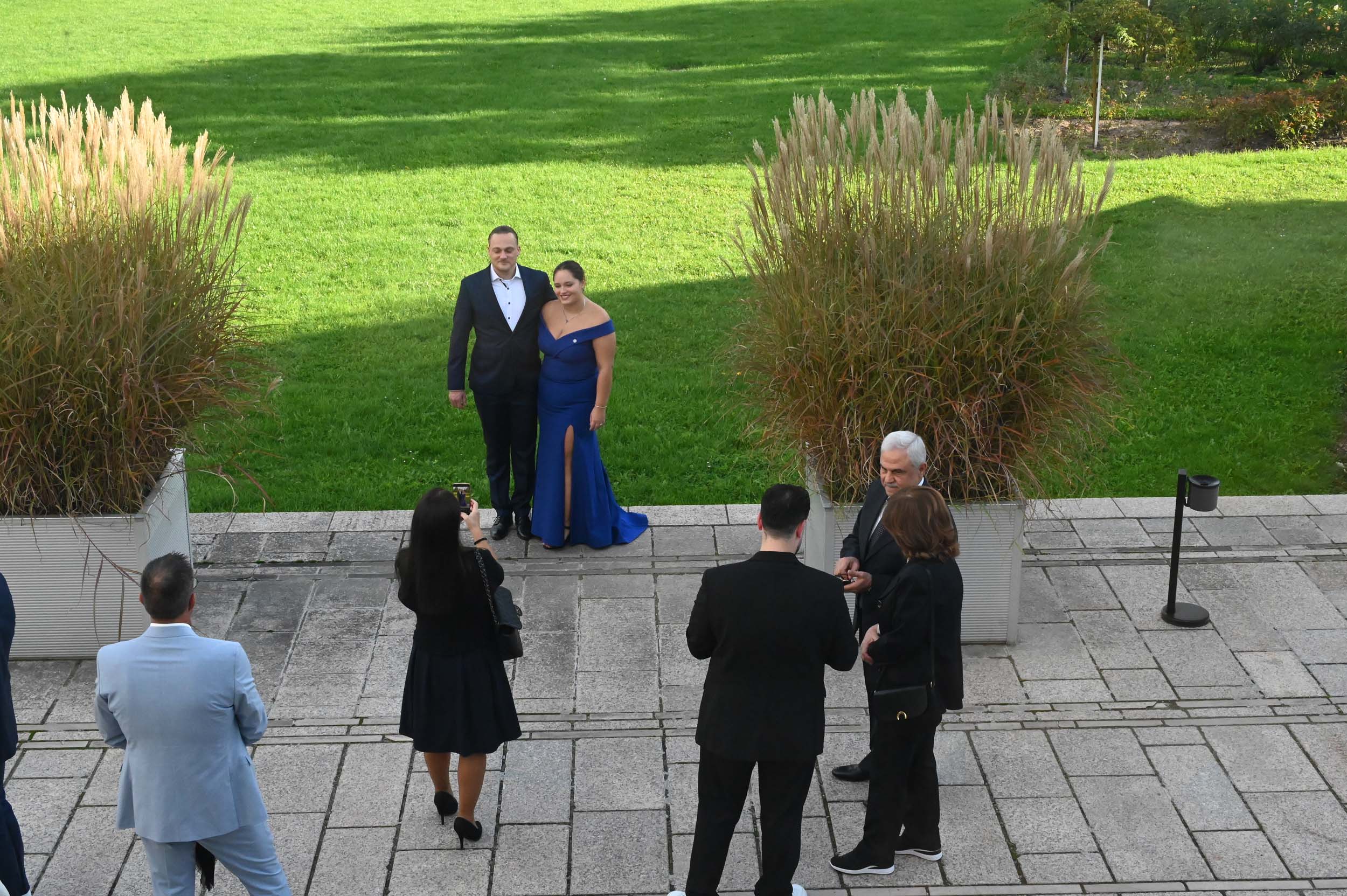 Ein Paar posiert für ein Foto auf einem Steinweg vor hohem Gras, während mehrere Absolventen und Absolventinnen an einem sonnigen Tag mit einer grünen Wiese im Hintergrund daneben stehen und so die Stimmung des Abschieds und des Feierns einfangen. at Hochschule Coburg