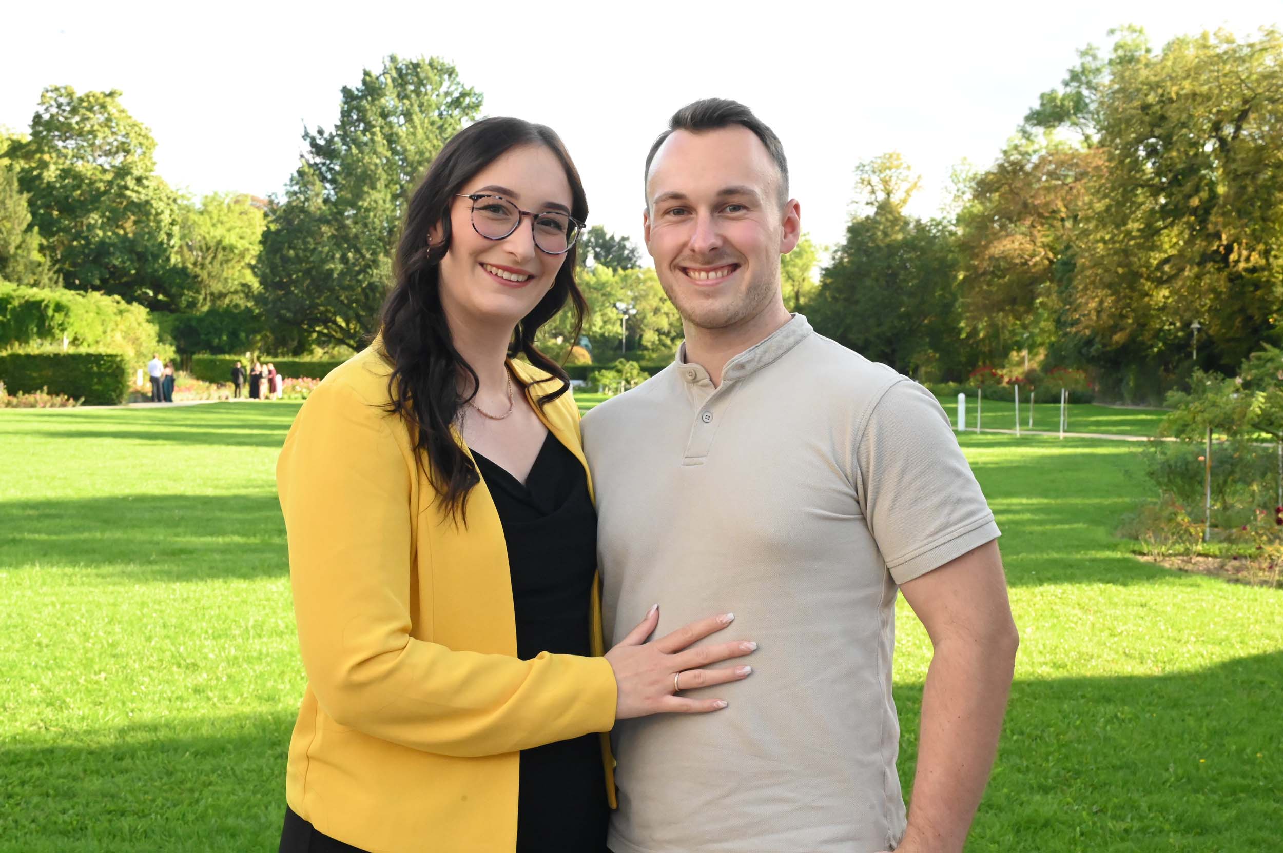 Eine Frau in einem gelben Blazer und ein Mann in einem hellen Poloshirt lächeln, während sie gemeinsam auf einer sonnigen Wiese mit Bäumen im Hintergrund stehen und als stolze Absolventen einen freudigen Abschiedsmoment festhalten. at Hochschule Coburg