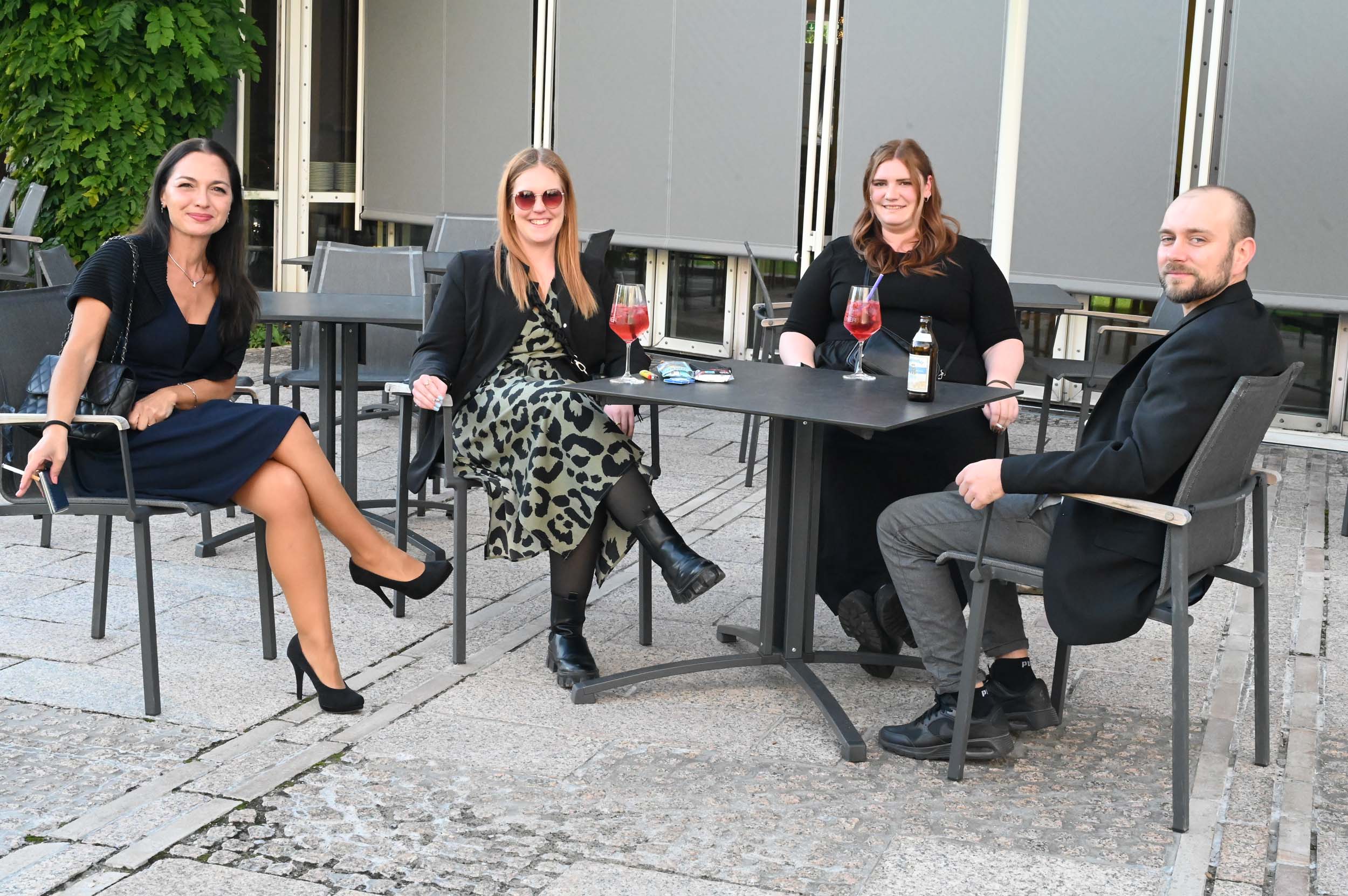 Vier Personen sitzen lächelnd an einem Tisch im Freien. Drei Frauen und ein Mann haben Getränke in der Hand. Diese Absolventen, die halbformelle Kleidung tragen, wirken entspannt und genießen ein ungezwungenes Abschiedstreffen auf einer Steinterrasse. at Hochschule Coburg