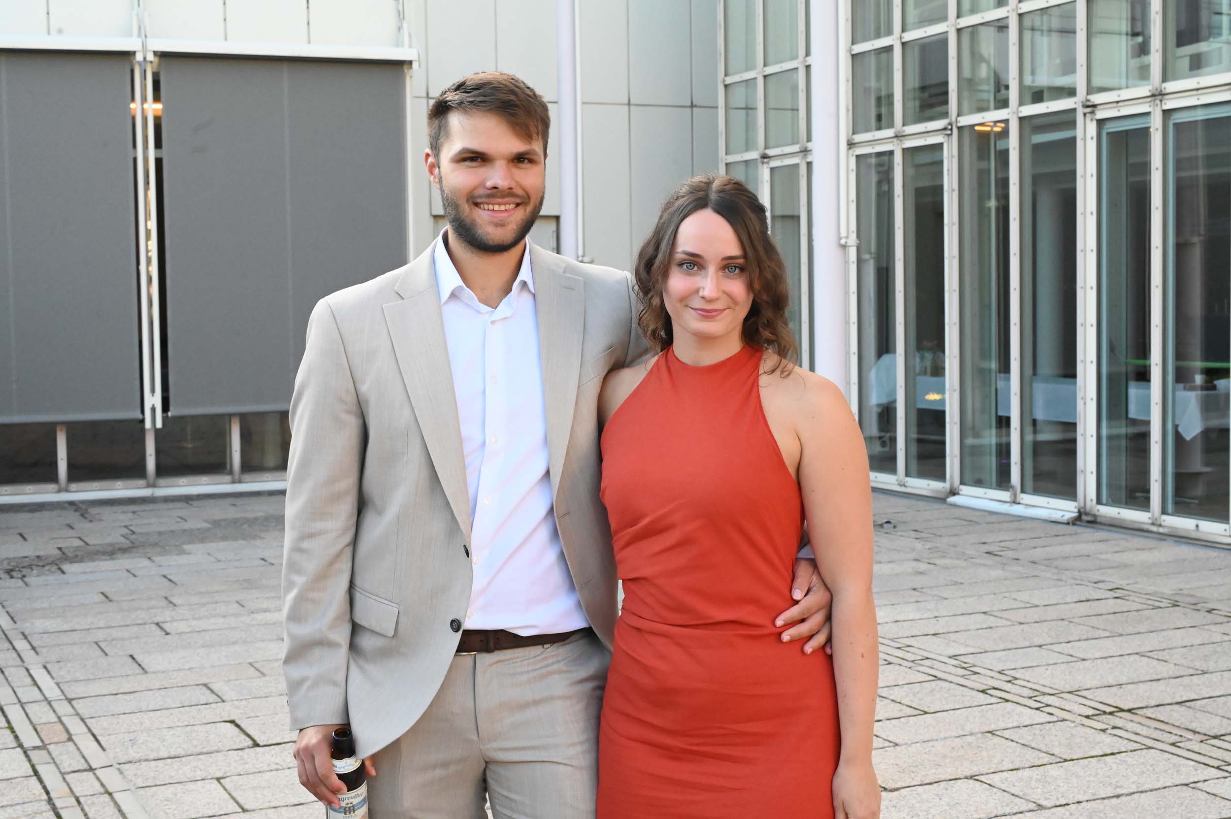 Ein Mann in einem hellen Anzug und eine Frau in einem roten, ärmellosen Kleid stehen gemeinsam vor einem modernen Glasgebäude und lächeln in die Kamera – ein fröhlicher Abschied, bei dem die Absolventen das Ende ihres Studiums feiern. at Hochschule Coburg