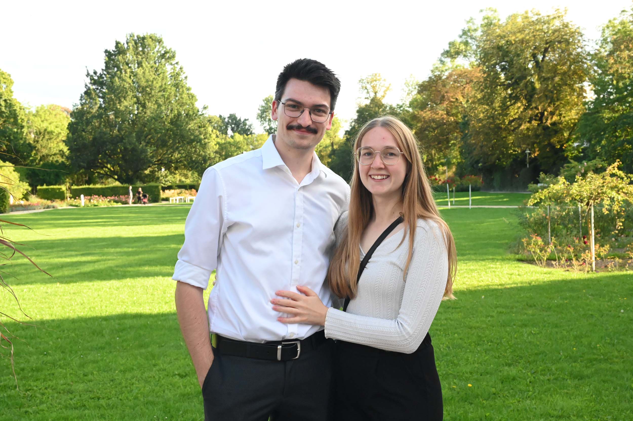 Ein junger Absolvent und eine junge Absolventin stehen dicht beieinander und lächeln in einem hellgrünen Park in die Kamera. Der Mann trägt eine Brille und ein weißes Hemd; die Frau hat lange Haare, eine Brille und einen hellen Pullover. Im Hintergrund sind Bäume und Gras zu sehen, die ihren Abschied vom Studentenleben einfangen. at Hochschule Coburg