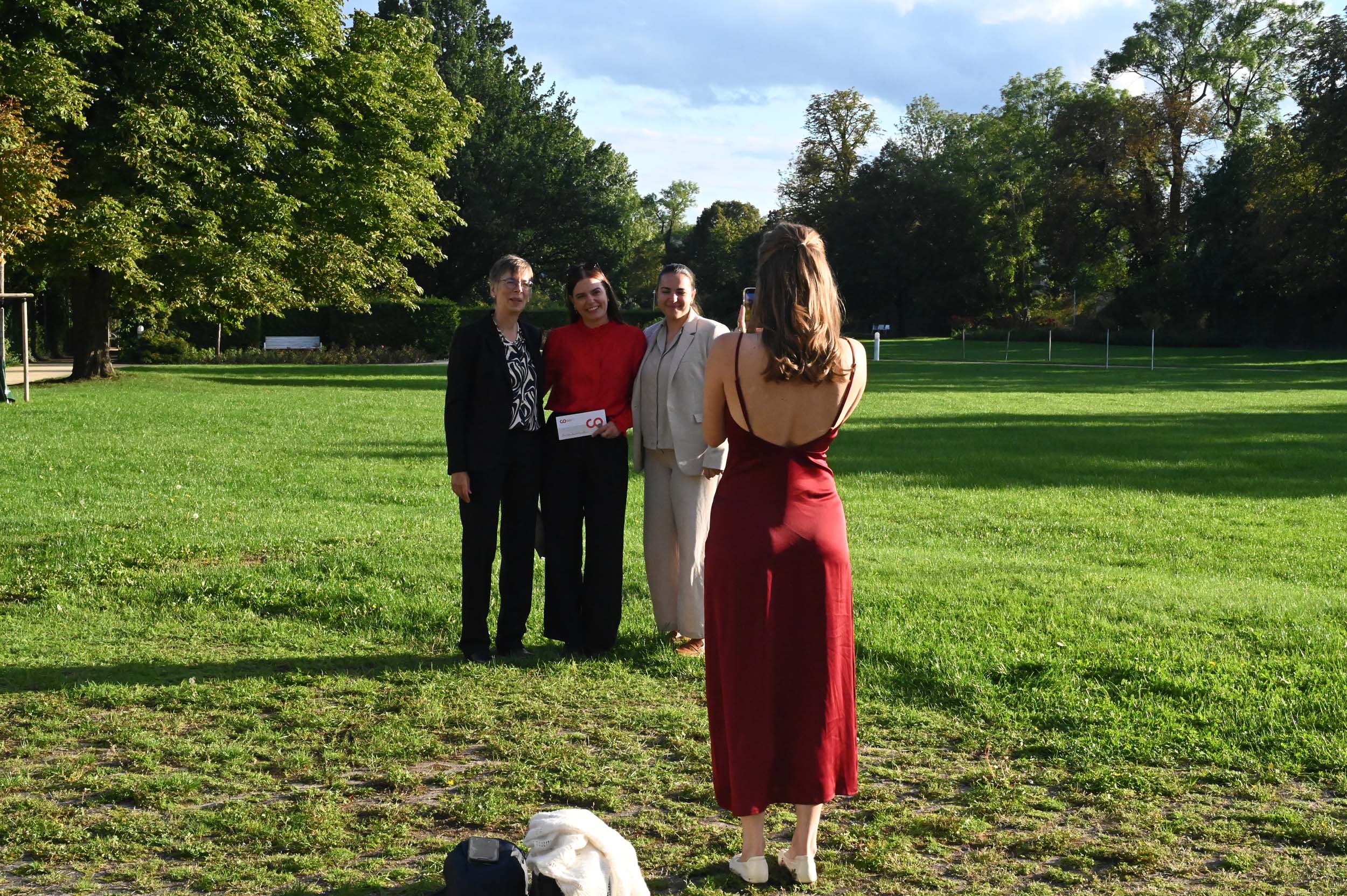 Vier Personen posieren auf einer Wiese für ein Foto; drei Absolventen stehen zusammen und blicken in die Kamera, während eine Absolventin in einem roten Kleid mit dem Rücken zur Kamera steht und das Foto macht – und so einen besonderen Abschiedsmoment festhält. at Hochschule Coburg