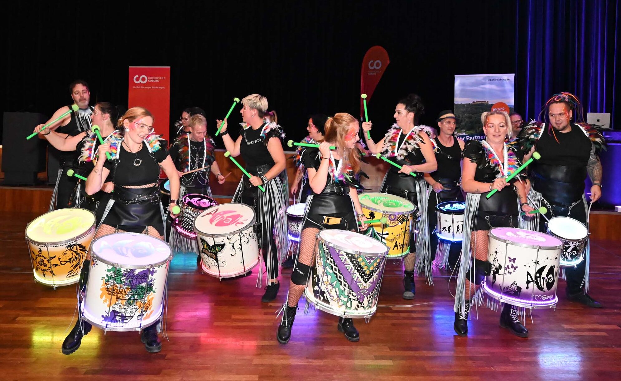 Eine Gruppe von Trommlern in schwarzen Outfits liefert unter strahlendem Bühnenlicht mit farbenfroh verzierten Trommeln und grünen Trommelstöcken eine energiegeladene Darbietung und sorgt so für einen unvergesslichen Abschied für die Absolventen und Absolventinnen. at Hochschule Coburg