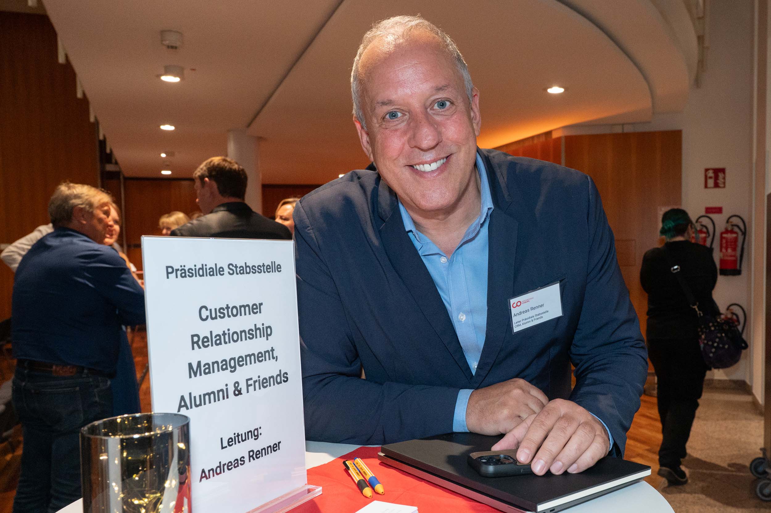 Ein lächelnder Mann in einem blauen Anzug steht an einem Schreibtisch mit der Aufschrift „Kundenbeziehungsmanagement, Alumni & Freunde“ und heißt Absolventinnen und Absolventen herzlich willkommen, während sich im Hintergrund bei einer Veranstaltung in den Innenräumen Menschen unterhalten. at Hochschule Coburg