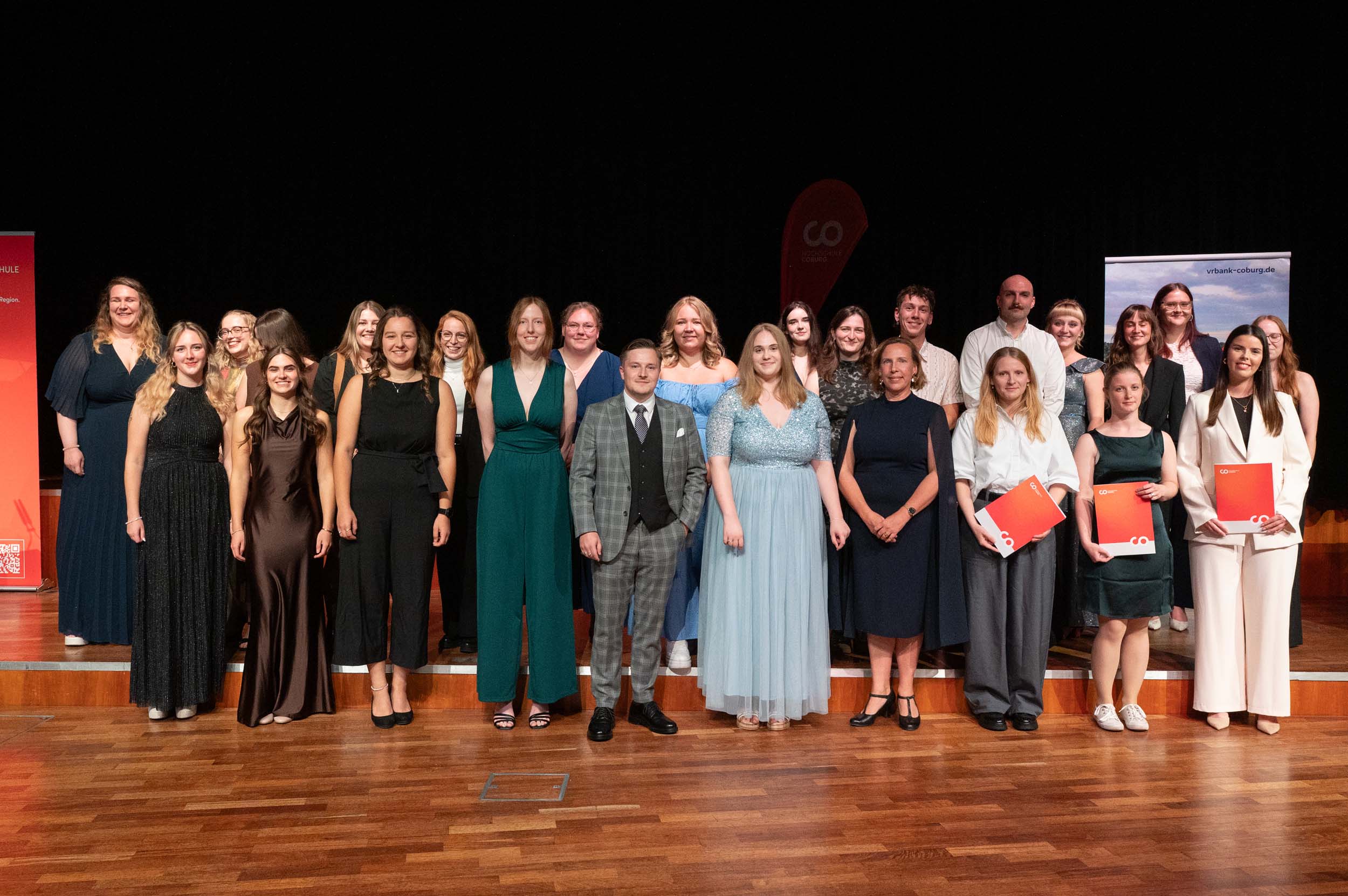Eine Gruppe von Absolventinnen und Absolventen in festlicher Kleidung steht für ein Gruppenfoto auf einer Bühne. Einige halten rote Mappen in den Händen, die ihren Abschied symbolisieren, während sie lächeln und in die Kamera blicken. Der Hintergrund ist dunkel, an beiden Seiten hängen Banner. at Hochschule Coburg
