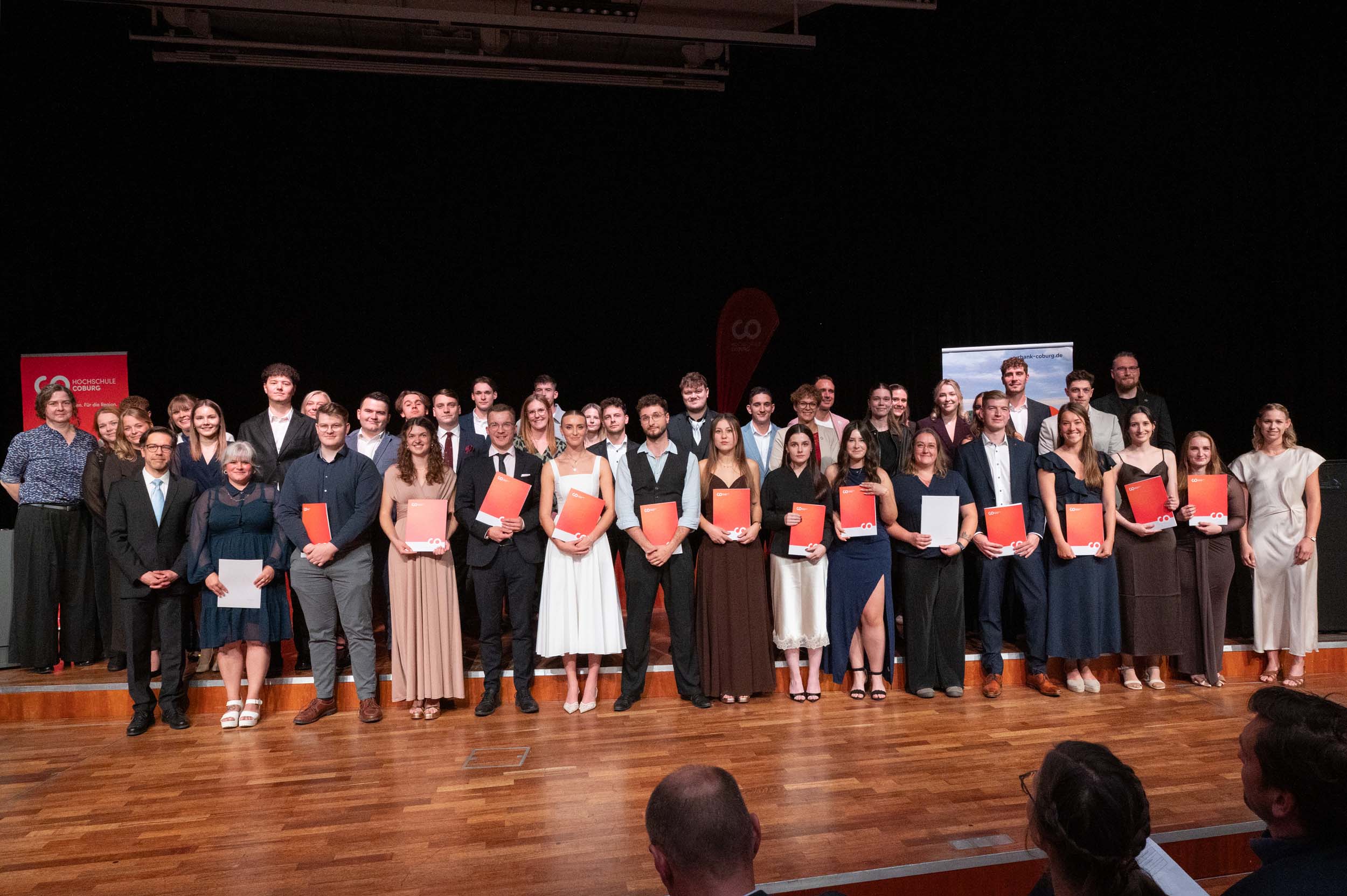 Eine große Gruppe von Menschen steht auf der Bühne, festlich gekleidet, mit Urkunden und Mappen in den Händen, und lächelt für ein Gruppenfoto. Der Hintergrund ist dunkel, und die Bühne hat einen Holzboden. Dieser feierliche Moment hält den Abschied der Absolventinnen und Absolventen fest, die den erfolgreichen Abschluss ihres Studiums feiern. at Hochschule Coburg