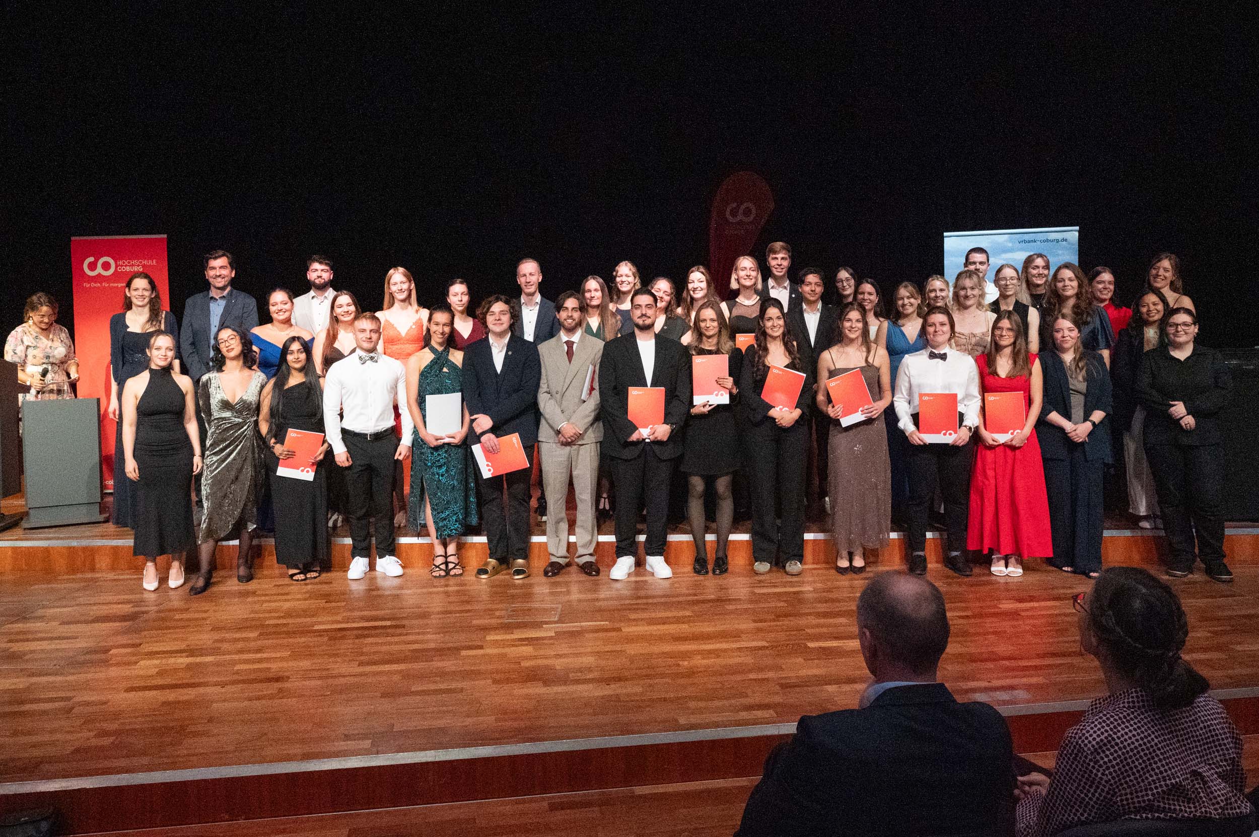 Eine große Gruppe von Absolventinnen und Absolventen in festlicher Kleidung steht auf einer Holzbühne, hält ihre Zeugnisse in den Händen und lächelt für ein Gruppenfoto, das ihren Abschied dokumentiert; im Vordergrund sind einige sitzende Zuschauer zu sehen. at Hochschule Coburg