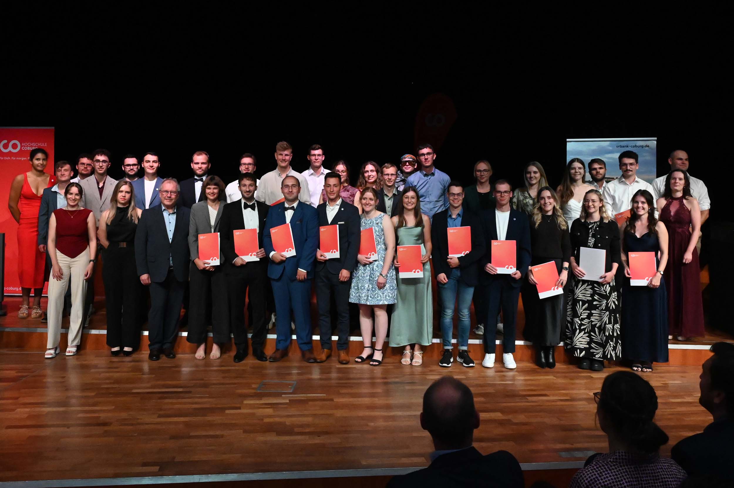 Eine große Gruppe von Absolventinnen und Absolventen in festlicher Kleidung steht auf der Bühne, einige halten ihre Urkunden in den Händen, und lächelt für ein Gruppenfoto. Der Hintergrund ist dunkel, ein Holzboden ist zu sehen, und an beiden Seiten sind einige Banner zu erkennen, die die Stimmung des Abschieds einfangen, während sie gemeinsam ihre Erfolge feiern. at Hochschule Coburg
