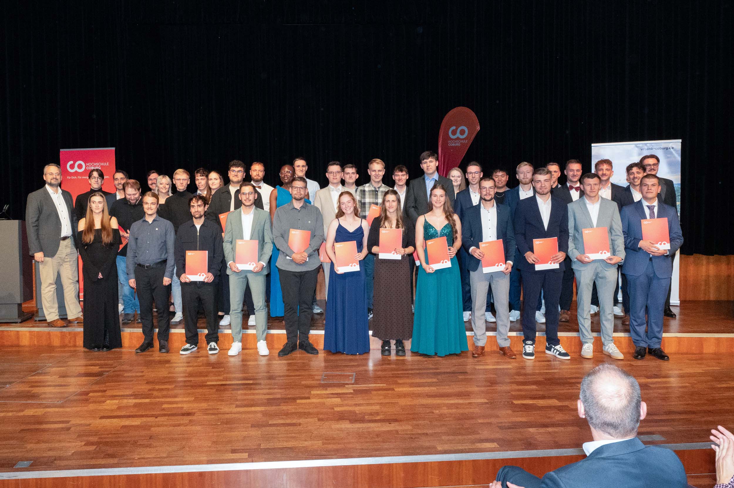 Eine große Gruppe von Menschen, darunter Absolventinnen und Absolventen, steht mit ihren Zeugnissen in der Hand auf einer Bühne und posiert für ein Foto. Die meisten sind festlich gekleidet, und im Hintergrund sind Transparente zu sehen. Im Vordergrund sitzt ein Zuschauer, der diesem unvergesslichen Abschiedsfest beiwohnt. at Hochschule Coburg