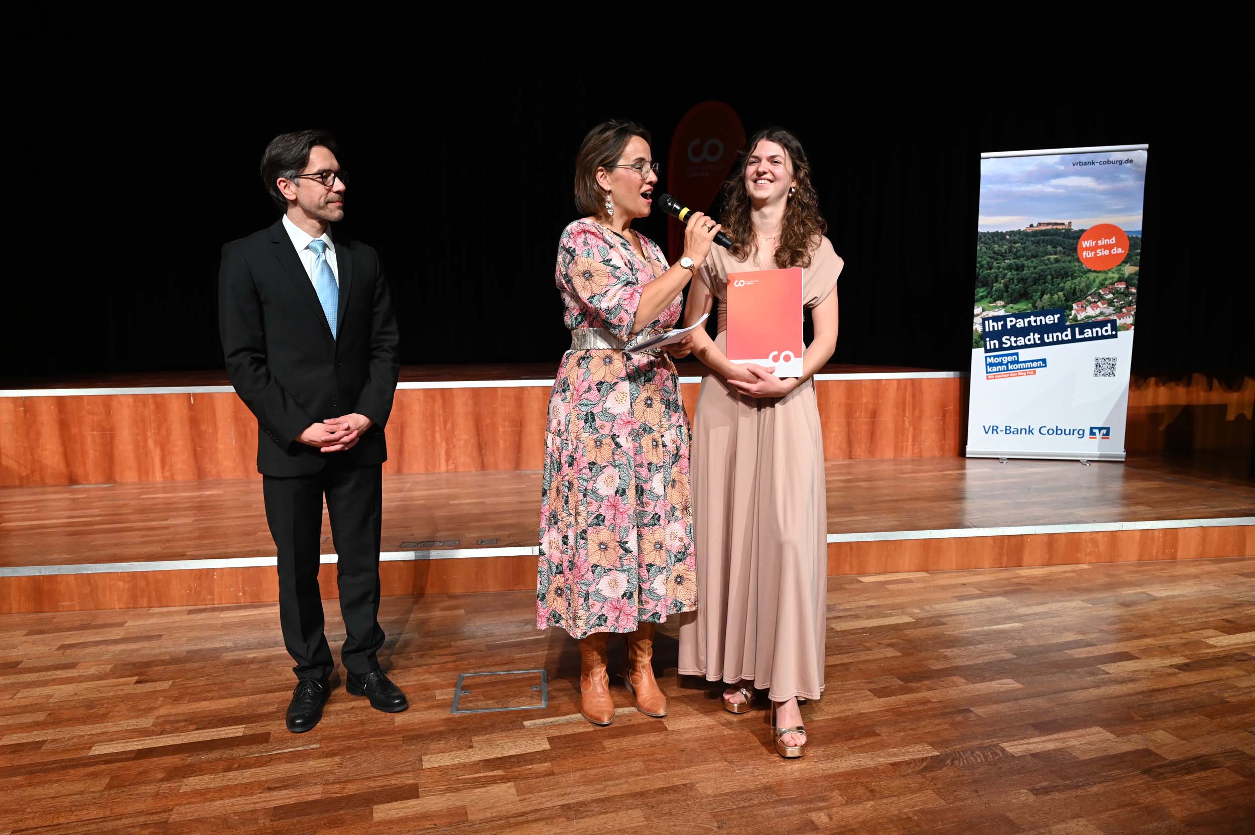 Drei Personen stehen auf einer Bühne. Eine Frau in einem geblümten Kleid spricht in ein Mikrofon und überreicht einer lächelnden Absolventin in einem beigen Kleid eine rote Mappe, während ein Mann im Anzug zusieht. Im Hintergrund steht ein Werbebanner, das den festlichen Abschied hervorhebt. at Hochschule Coburg