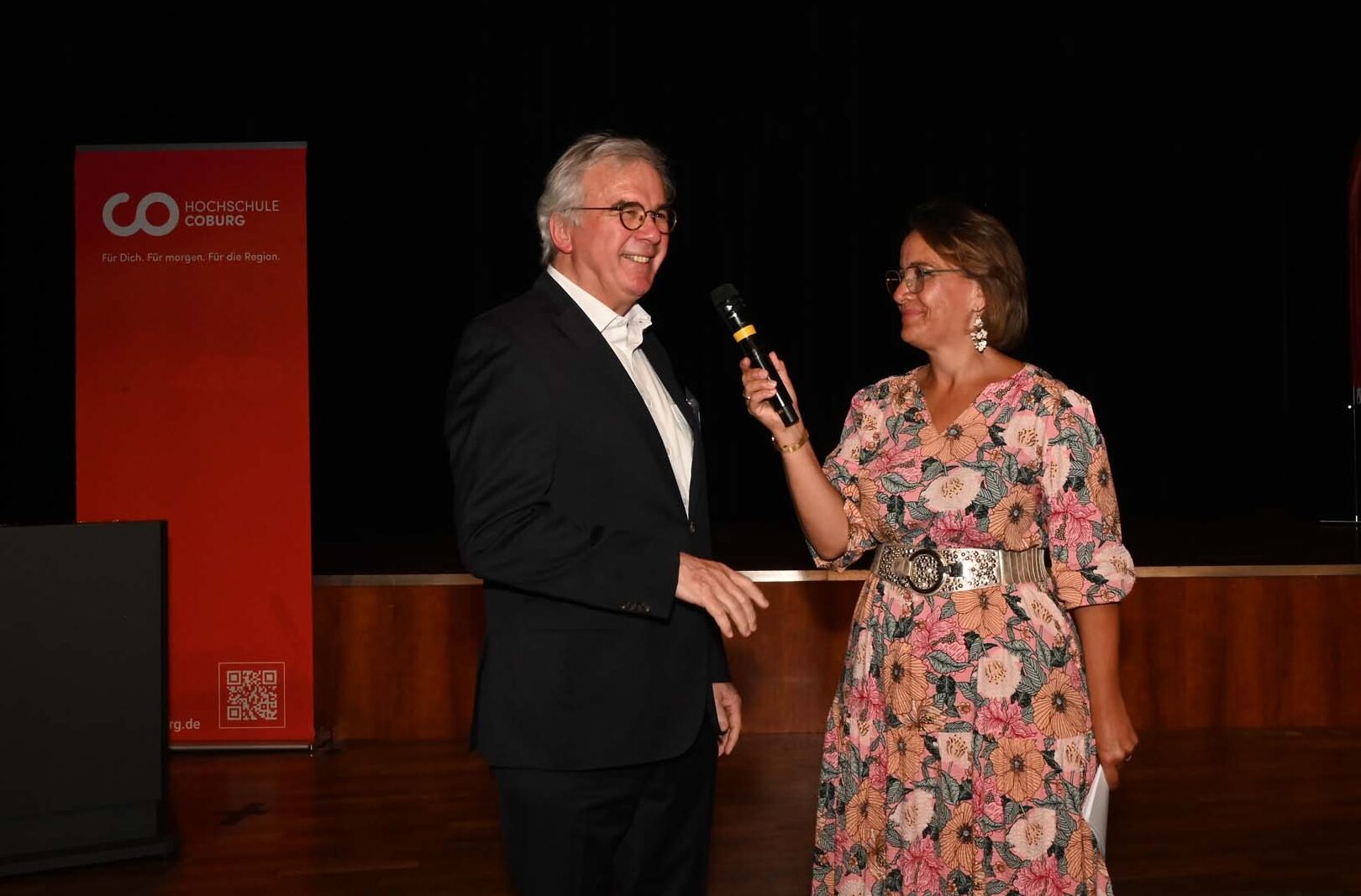 Eine Frau in einem geblümten Kleid hält ein Mikrofon in der Hand und interviewt einen Mann im Anzug auf einer Bühne mit Holzboden und roten Transparenten im Hintergrund, während sie den Abschied der diesjährigen Absolventinnen und Absolventen feiern. at Hochschule Coburg