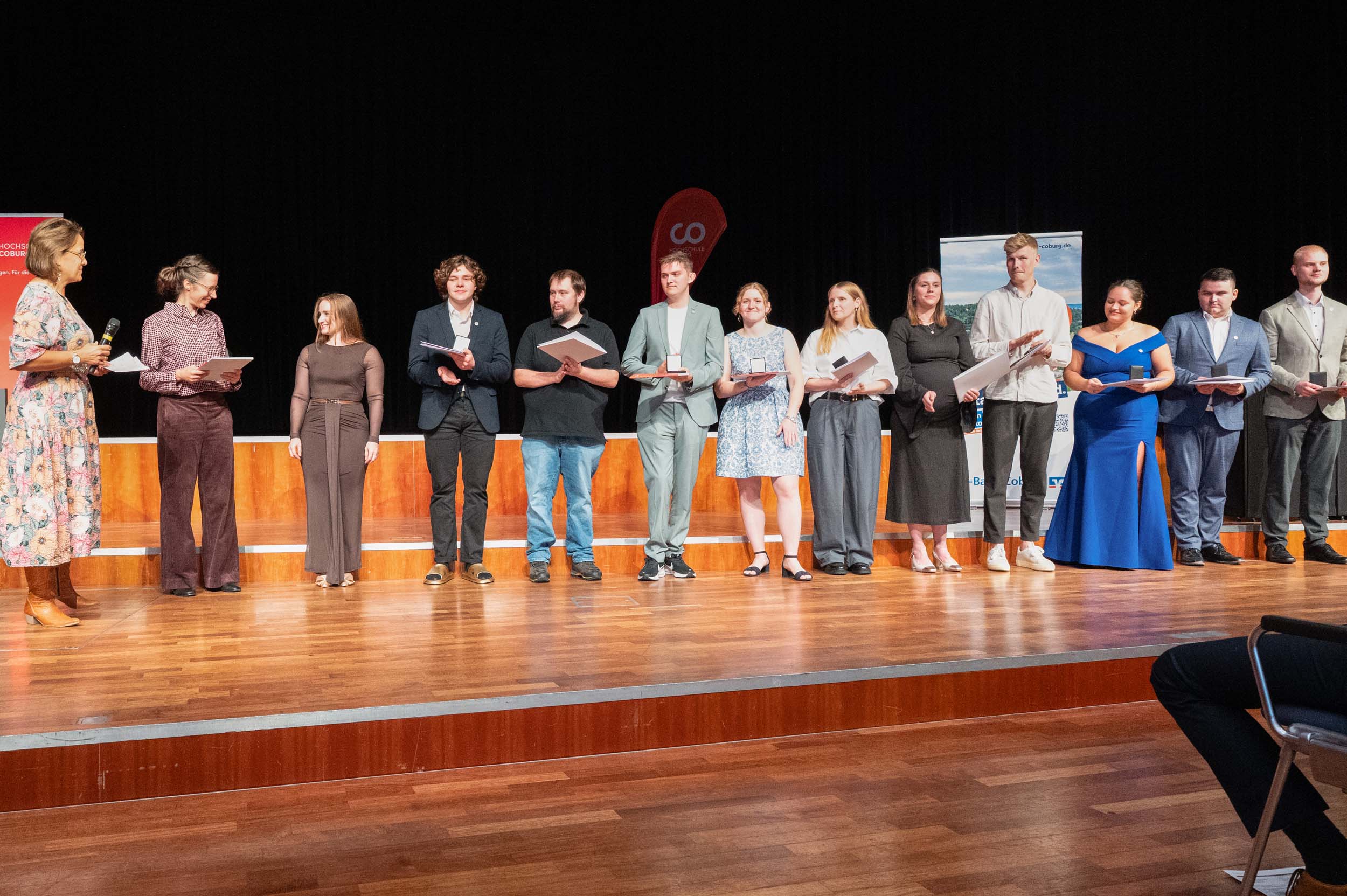 Eine Gruppe von Absolventen und Absolventinnen steht in einer Reihe auf einer Bühne und hält ihre Zeugnisse in den Händen. Sie blicken zum Publikum, sind halbformell gekleidet, und links scheint eine Frau in diesem Moment des Abschieds eine Ansprache an sie zu richten. Der Hintergrund ist dunkel, wodurch die Teilnehmer besonders hervorgehoben werden. at Hochschule Coburg