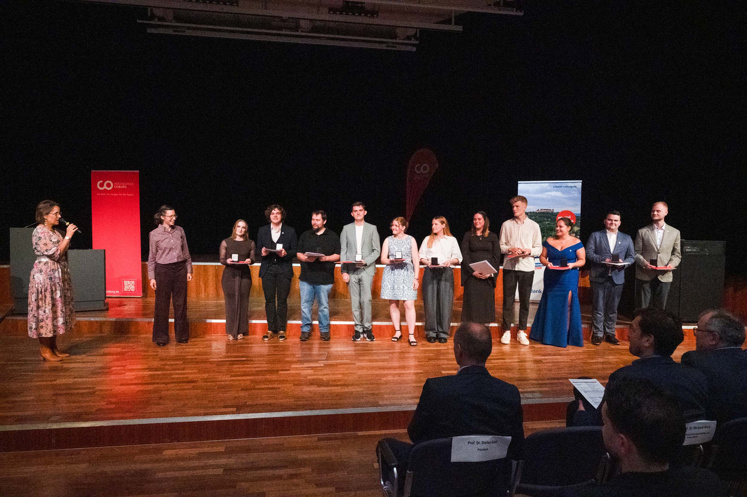Eine Gruppe von Absolventen und Absolventinnen steht mit ihren Abschlusszeugnissen auf der Bühne, während eine Frau mit einem Mikrofon zu ihnen spricht. Im Vordergrund sitzen Zuschauer, und im Hintergrund sind Transparente zu sehen, die die Bedeutung dieses Abschiedsmoments unterstreichen. at Hochschule Coburg