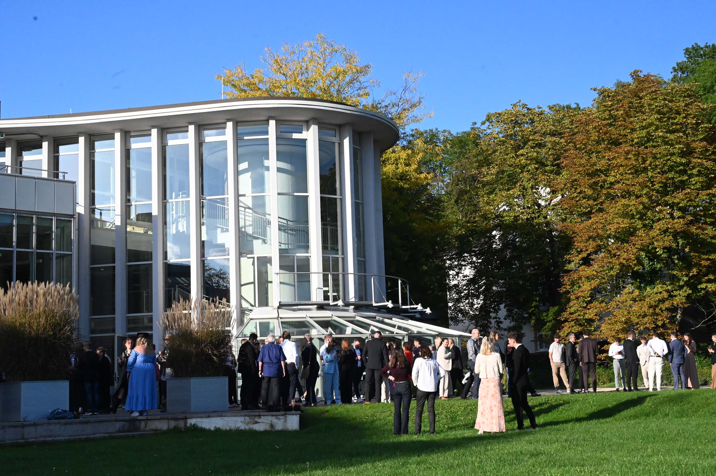 An einem sonnigen Tag versammeln sich Menschen in festlicher Kleidung vor einem modernen Glasgebäude, während sich Absolventinnen und Absolventen zu einem emotionalen Abschied zusammenfinden – vor dem Hintergrund von grünem Gras und Bäumen mit Herbstlaub. at Hochschule Coburg